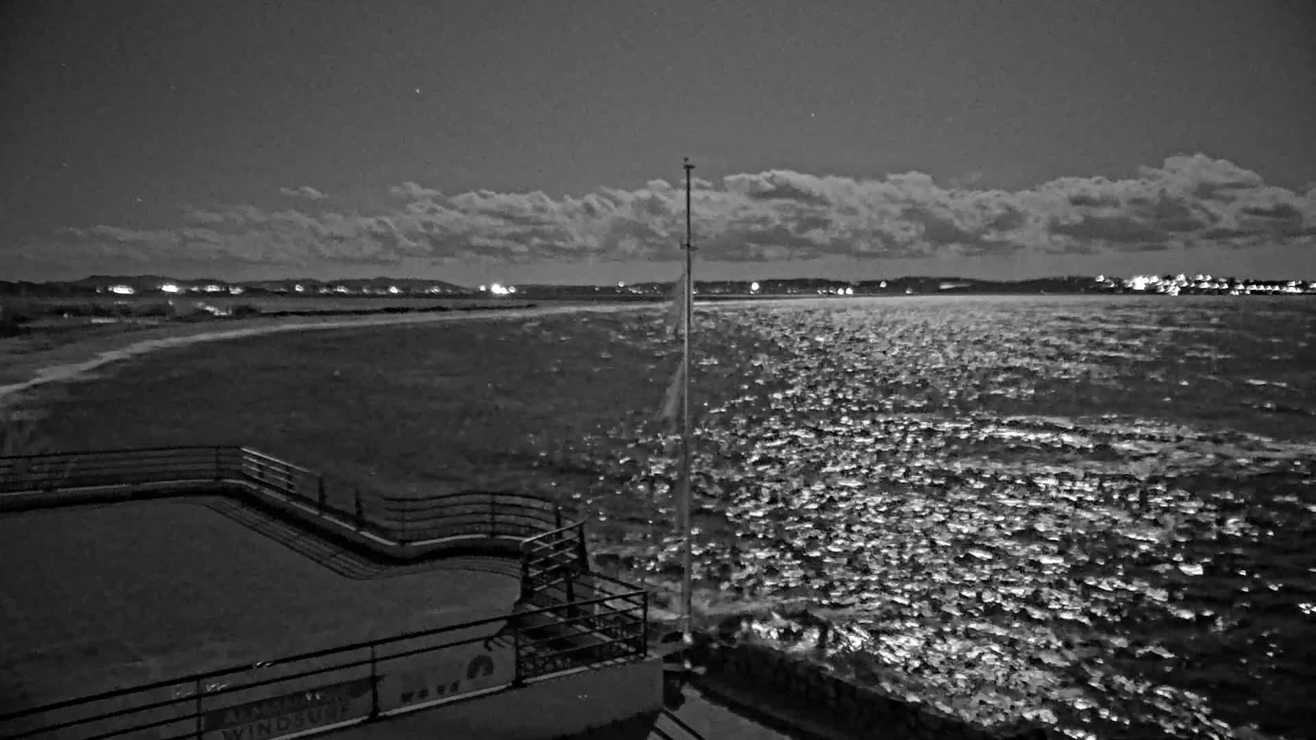 A nighttime, black and white view of a choppy body of water under a cloudy sky, with a distant shoreline showing lights and a railing at the foreground.