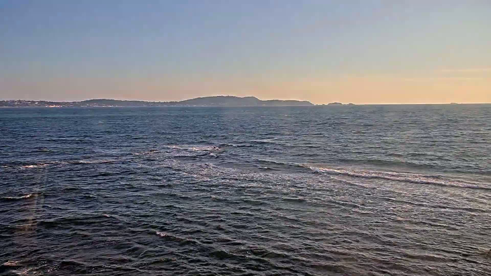 A calm ocean with small waves stretches to a distant landmass under a clear, light-blue sky.