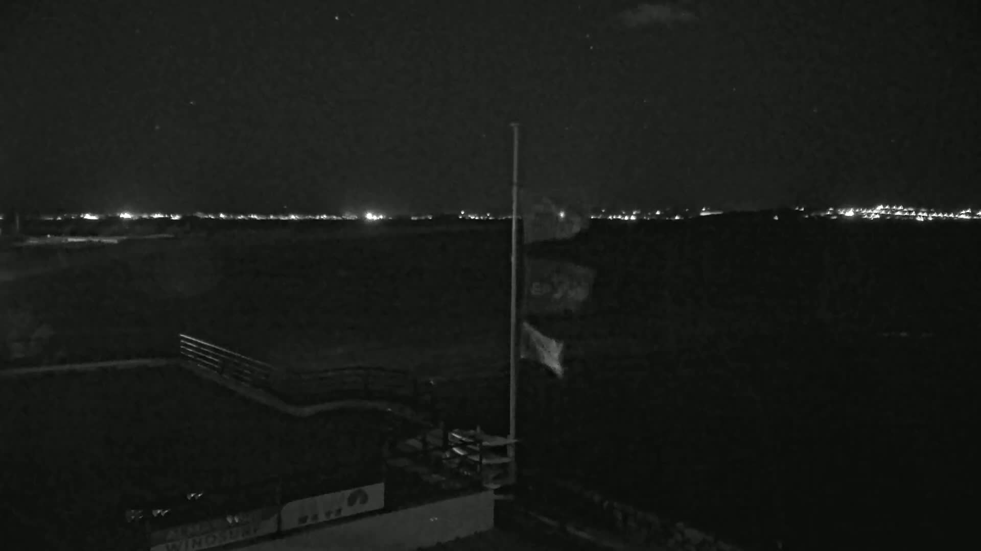 A nighttime, low-light view shows a flagpole with a flag, a railing, and a distant city skyline illuminated by lights under a clear, starry sky.
