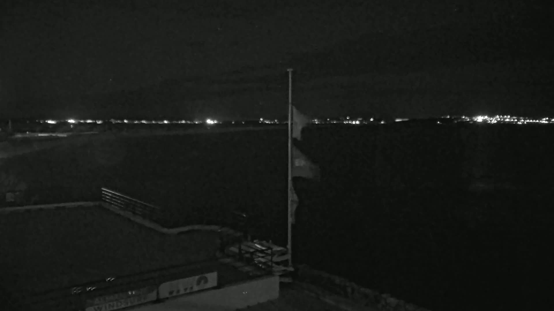 A nighttime view of a dark landscape with a flagpole and a distant line of city lights under a clear sky.