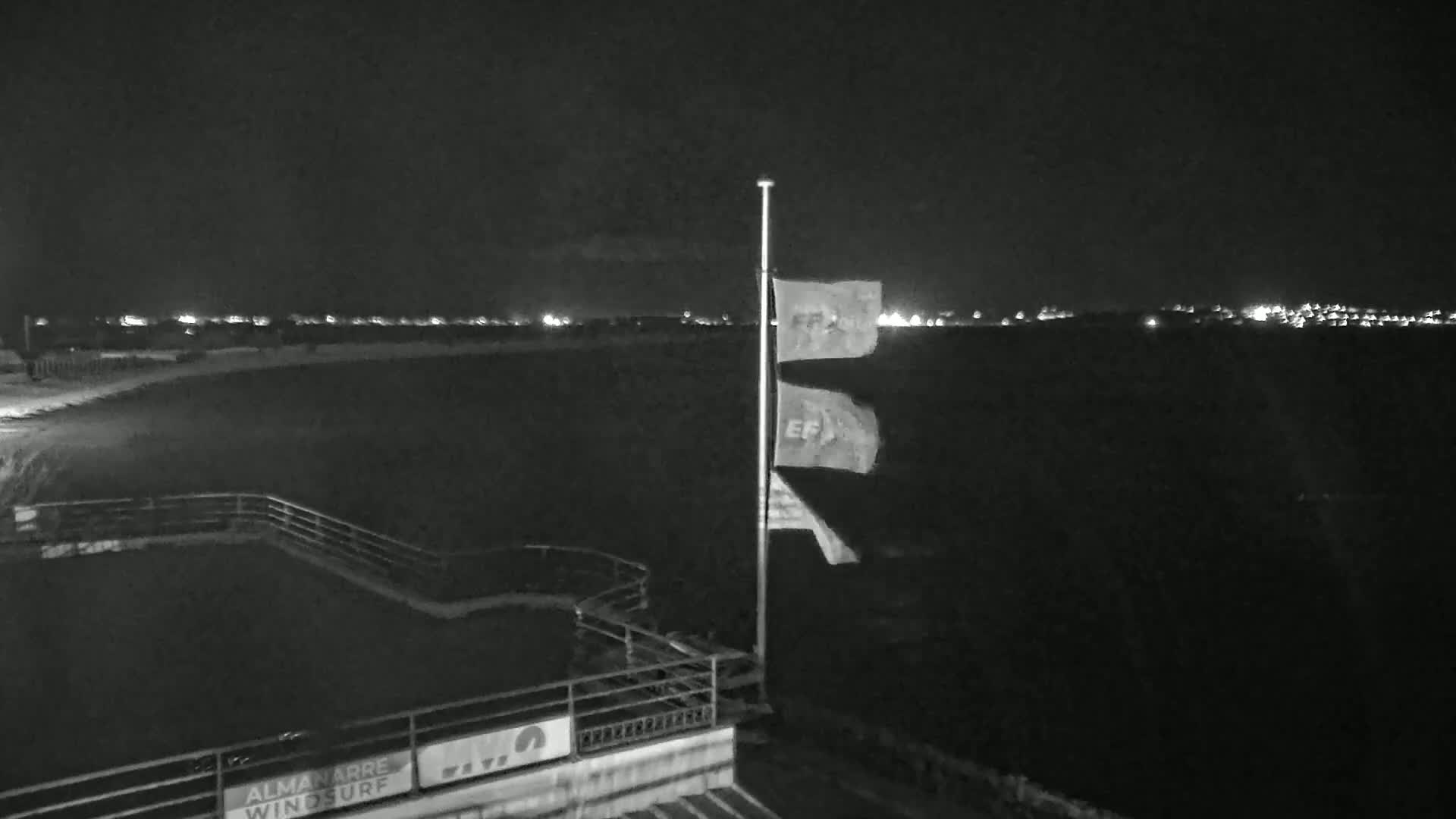 A nighttime view of a dark beach with a flagpole displaying three flags, and a distant line of lights on the horizon.
