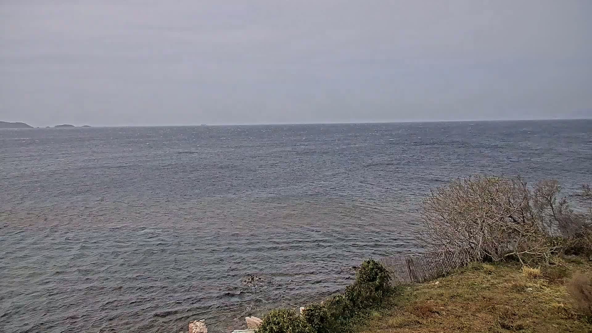 A choppy, dark-blue sea extends to a hazy horizon under a gray sky, with brown, leafless shrubs and scrubland visible in the foreground.