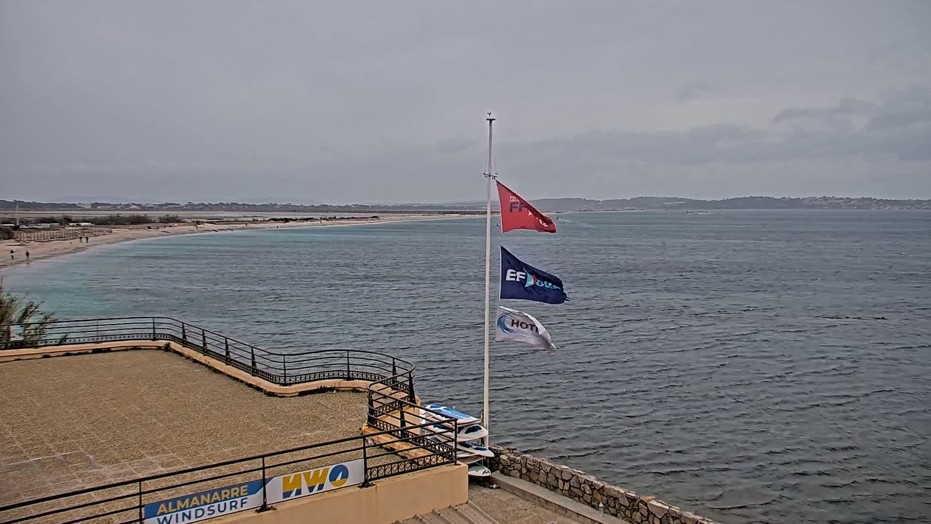 A flagpole with three flags stands on a paved area overlooking a calm, gray ocean and a sandy beach with a few people visible in the distance under an overcast sky.