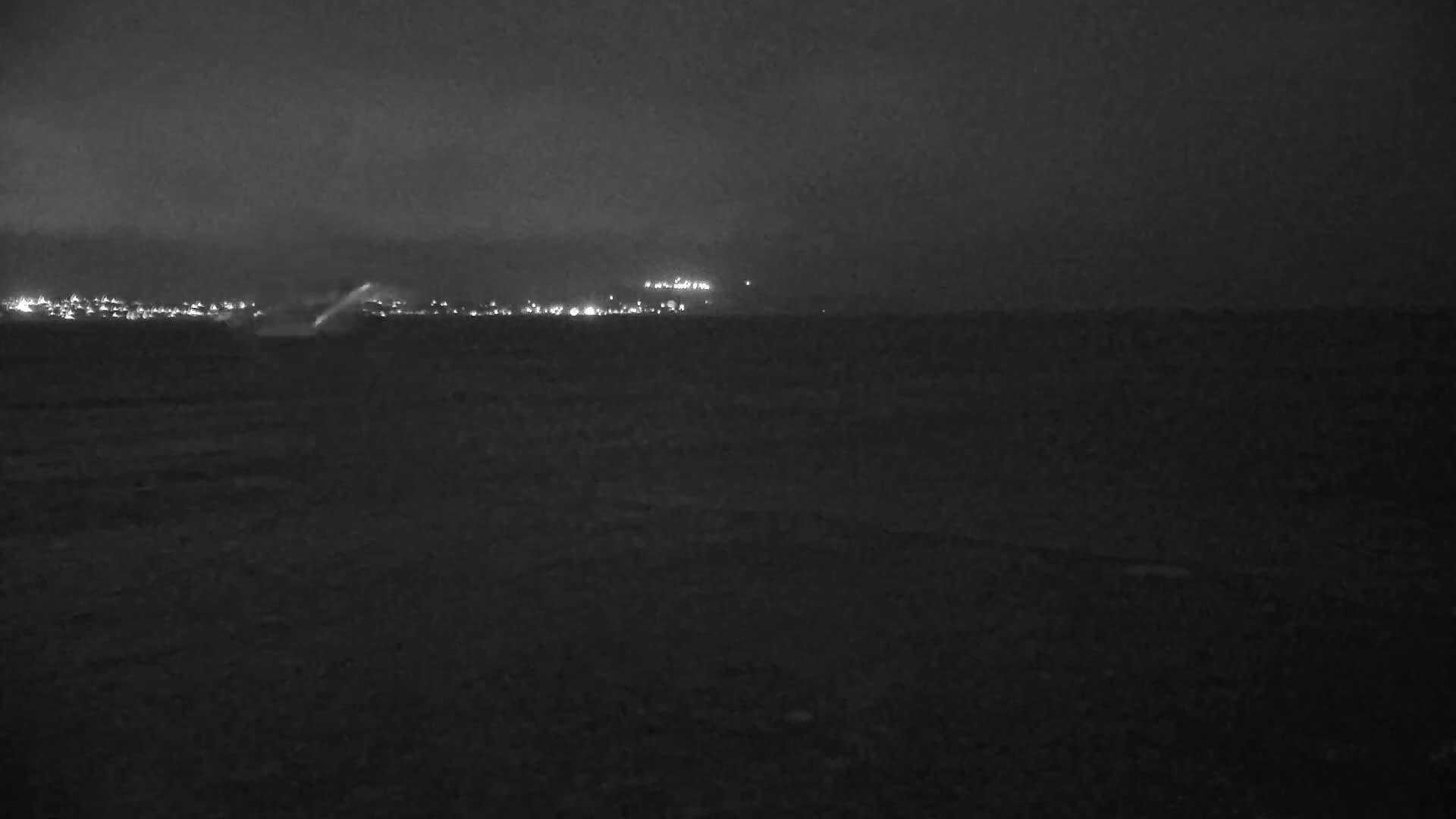 A dark, nighttime scene shows a distant line of city lights across a dark body of water under an overcast sky.