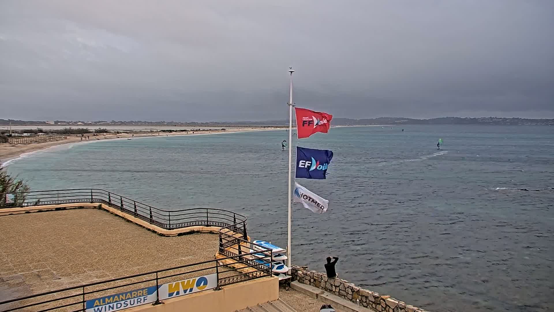 A beach with calm, light-blue water and overcast skies is viewed from an elevated vantage point, showing several windsurfers on the water and several flags on a pole near a stone wall.