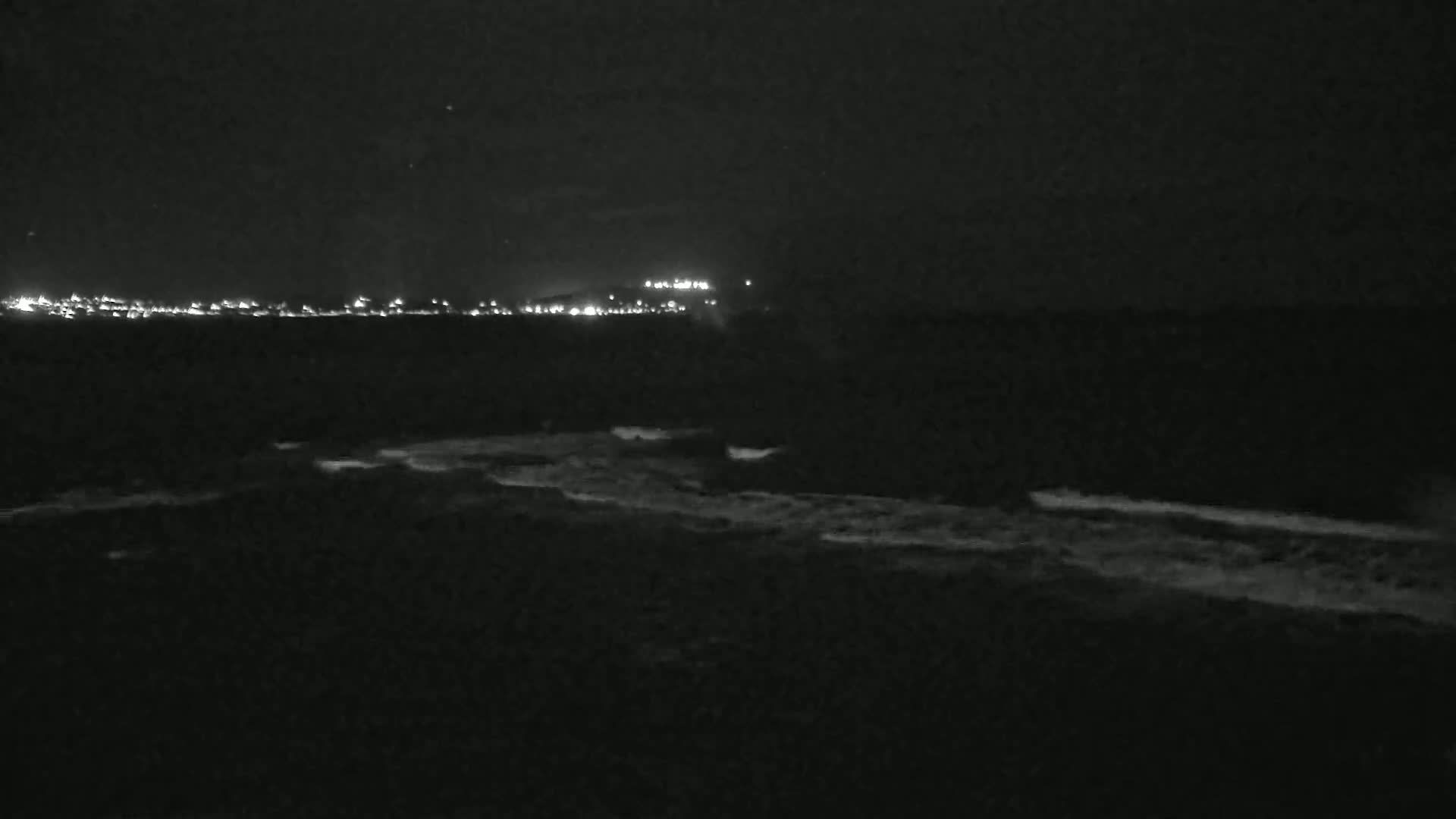 A dark, nighttime coastal scene shows a distant line of lights across the water, with gently breaking waves along the shoreline.