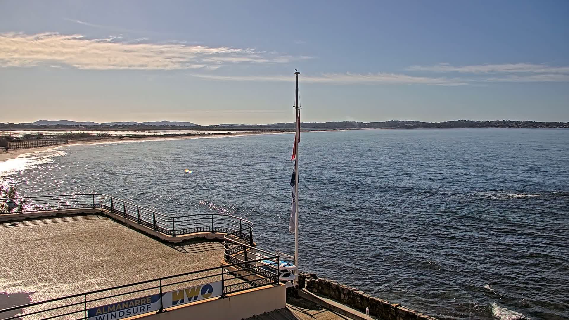 A mostly sunny day reveals a calm sea with a sandy beach and distant hills visible from a rooftop terrace with a flagpole.