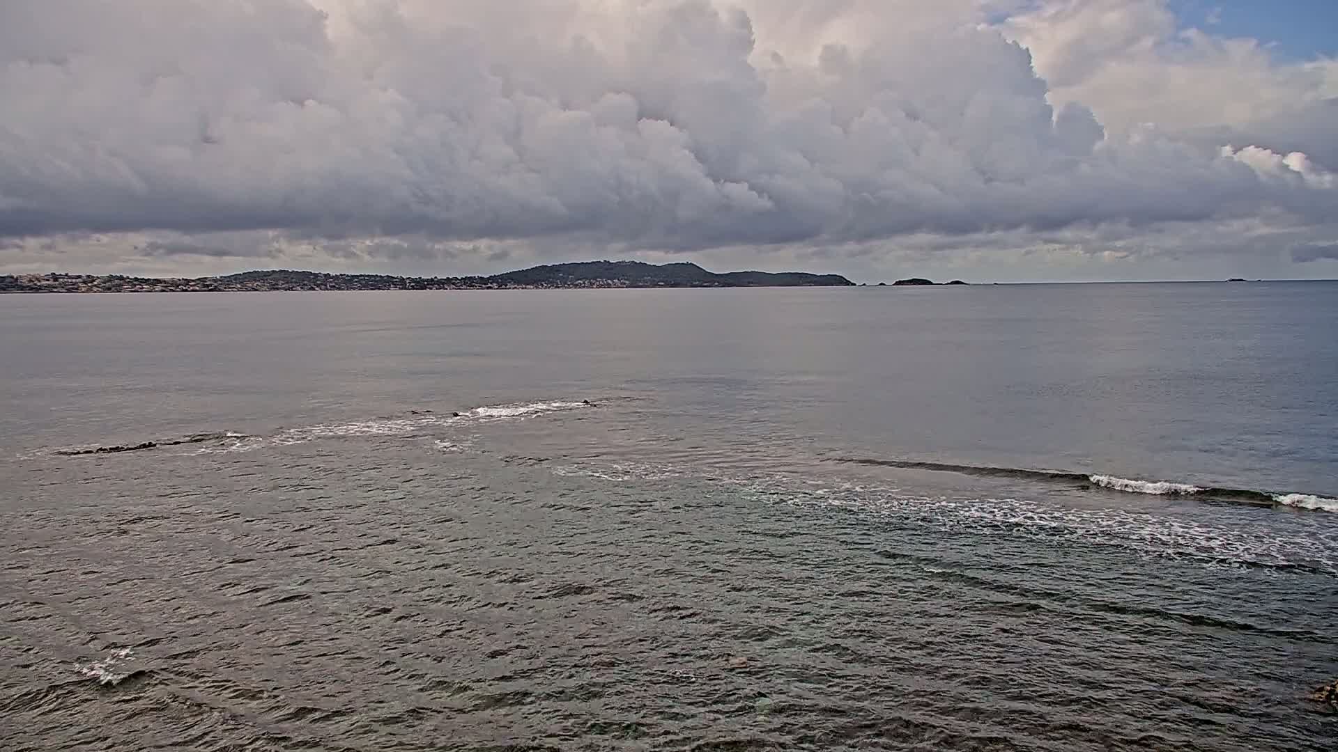 A calm sea with small waves laps against a rocky shore under a cloudy sky, with a distant landmass visible on the horizon.