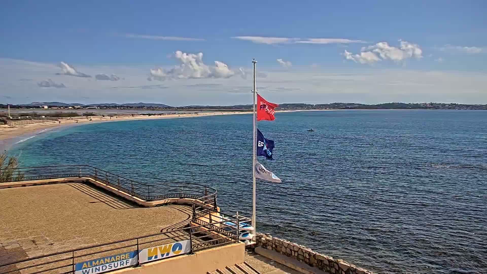A sandy beach curves along a calm, blue ocean under a mostly sunny sky with a few scattered clouds; a flagpole with three flags stands on a stone structure overlooking the water.