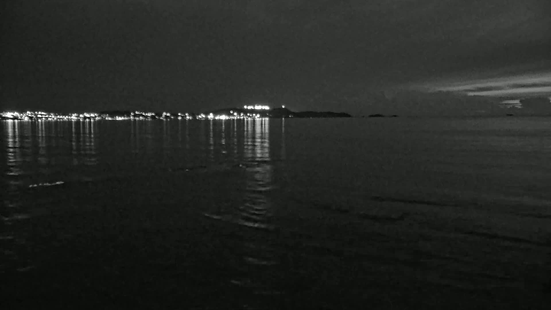 A nighttime view of a calm body of water reflecting the lights of a distant town on a dark, overcast night.