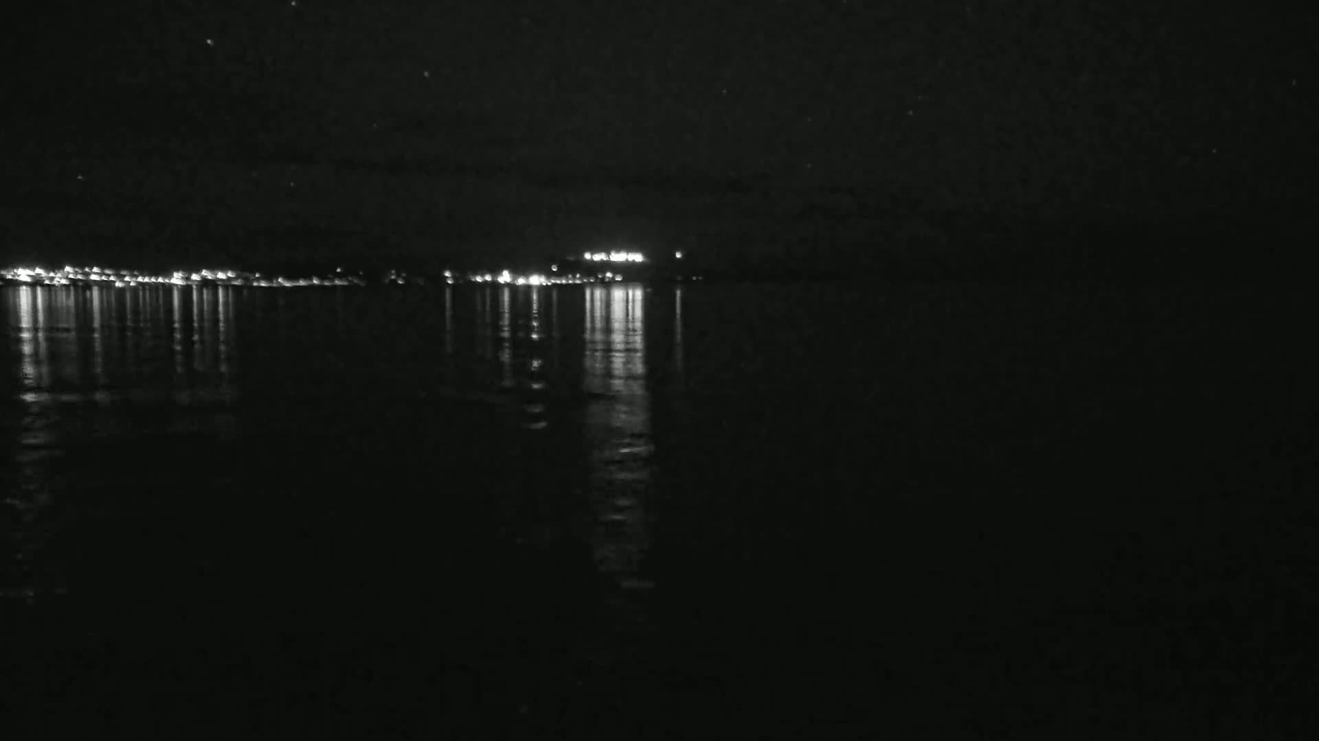A dark, nighttime scene shows city lights reflected on a calm body of water under a clear, starry sky.
