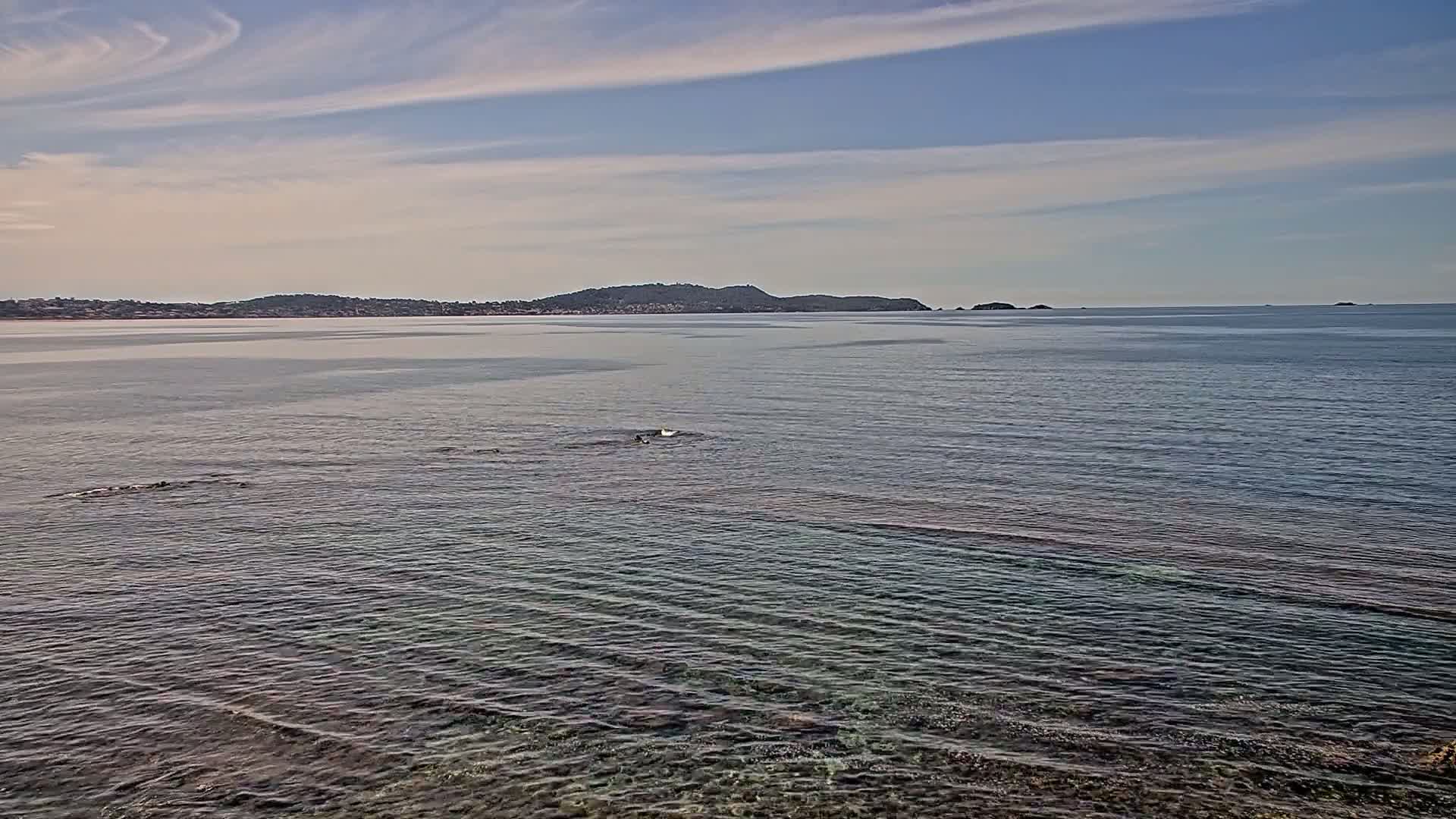 Calm, clear water laps gently at a rocky shore, with a distant landmass visible under a mostly clear sky.