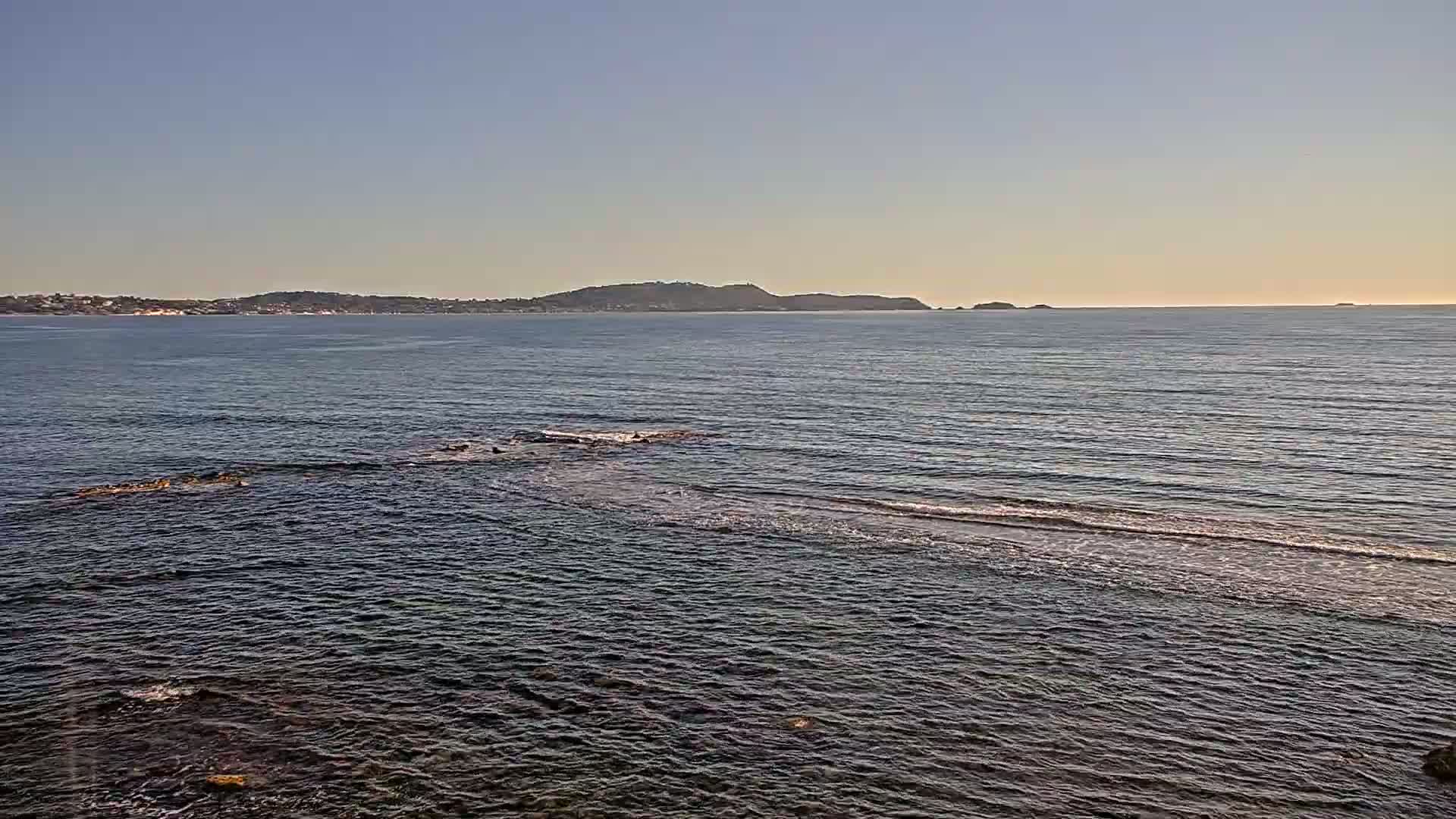A calm sea with small waves laps against dark rocks in the foreground, while a distant shoreline and islands are visible under a clear sky.