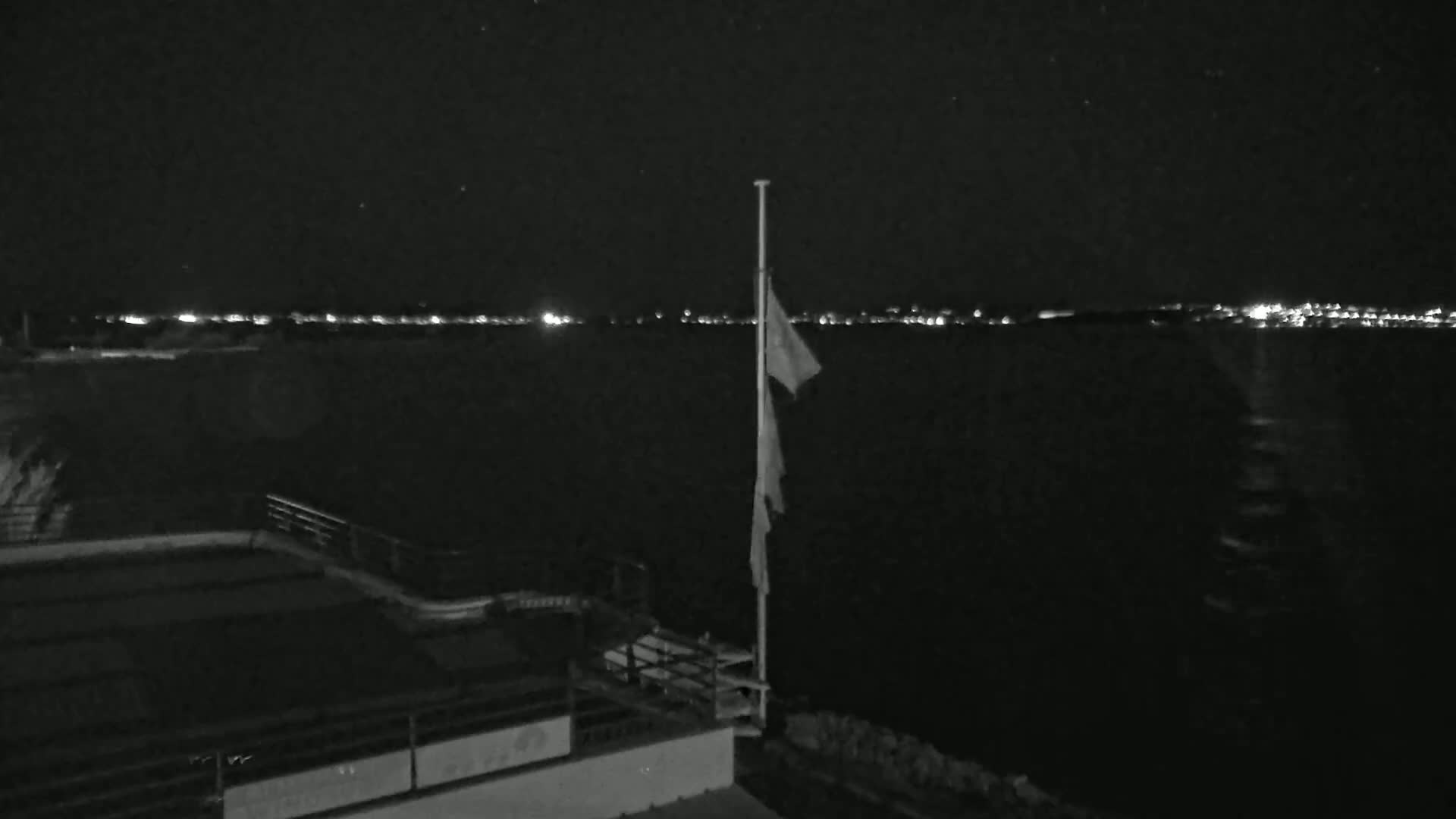 A nighttime view of a dark body of water with a flagpole and a tattered flag, a section of a building with a railing, and a distant shoreline with lights visible in the background under a clear, starry sky.