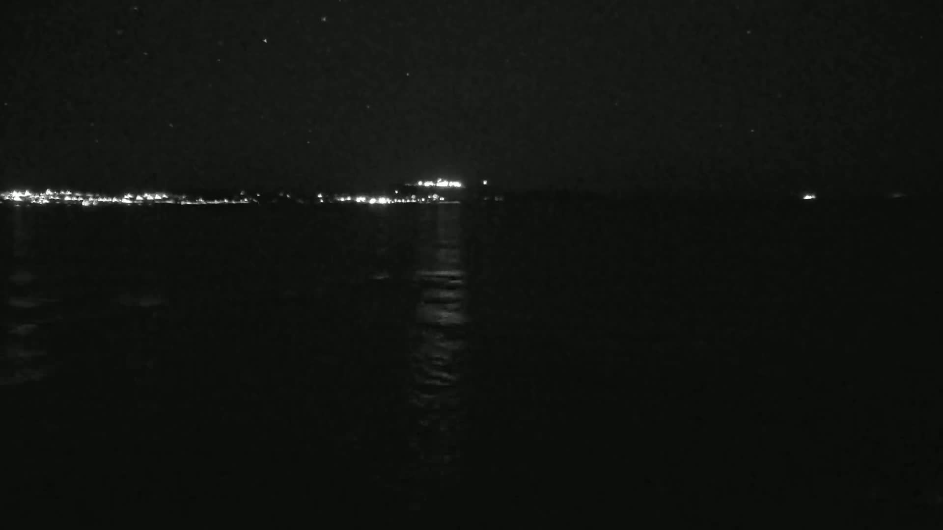 A nighttime view of a calm body of water reflecting light from a distant, faintly lit shoreline under a clear, starry sky.