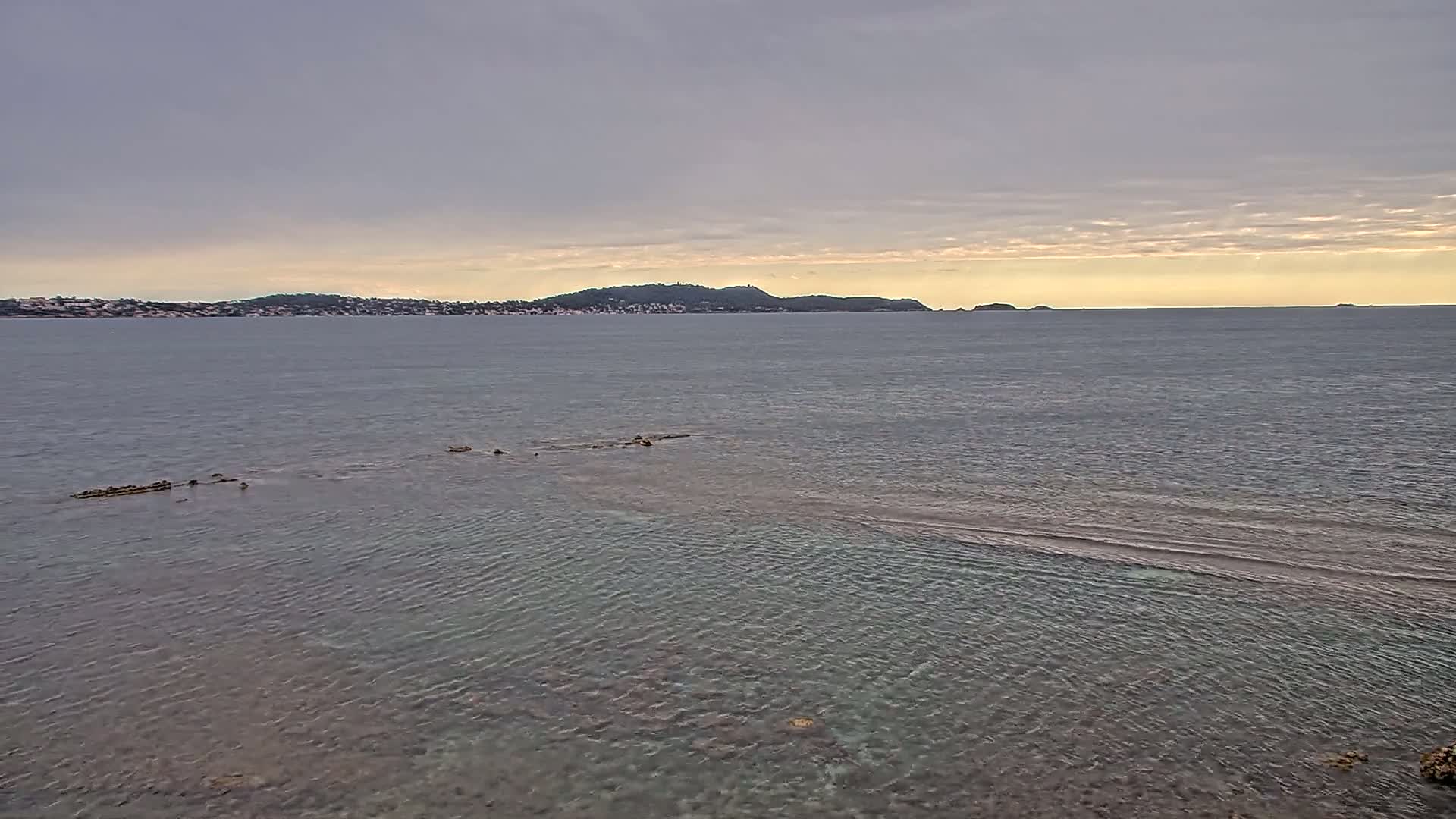 A calm sea with a partially submerged rock formation in the foreground, and a distant coastline under an overcast sky.