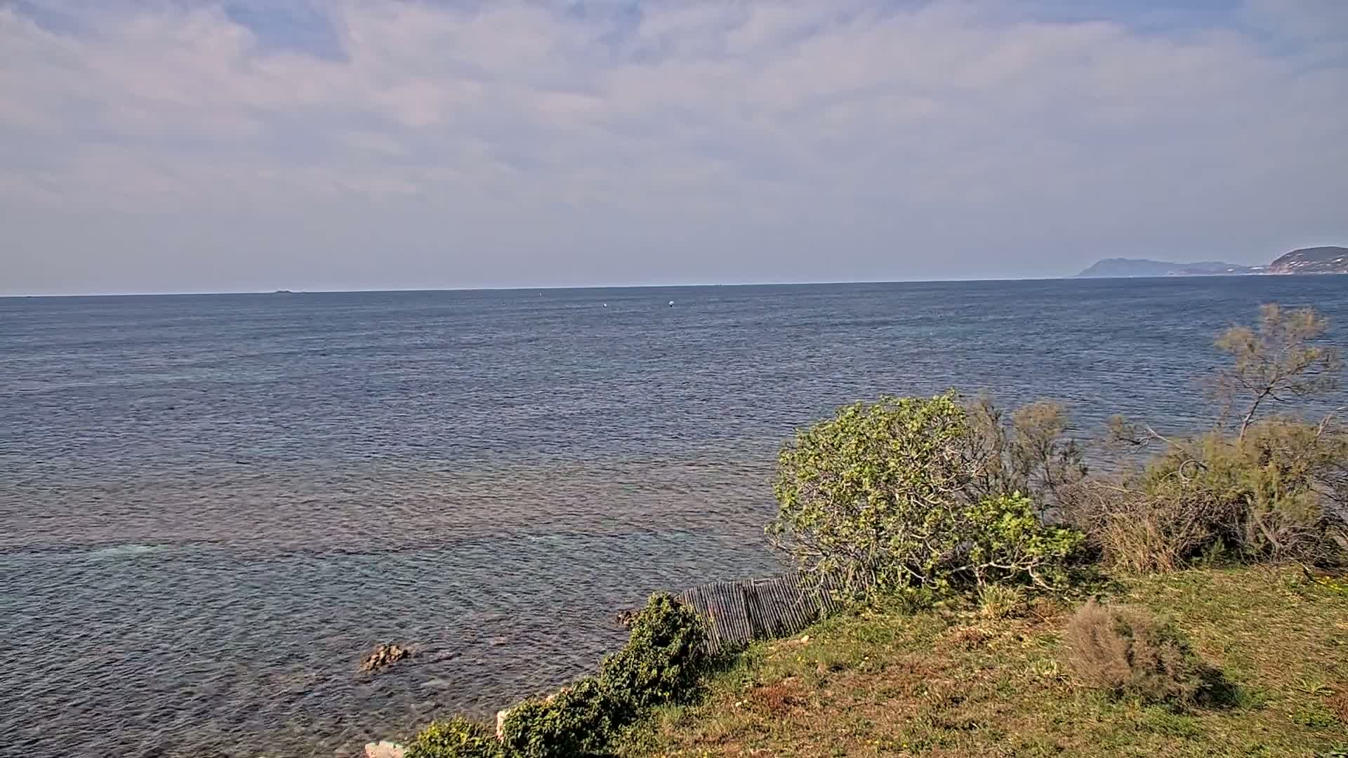 A partly cloudy day reveals a calm expanse of sea meeting a rocky shoreline, where scrubby vegetation grows along the edge.