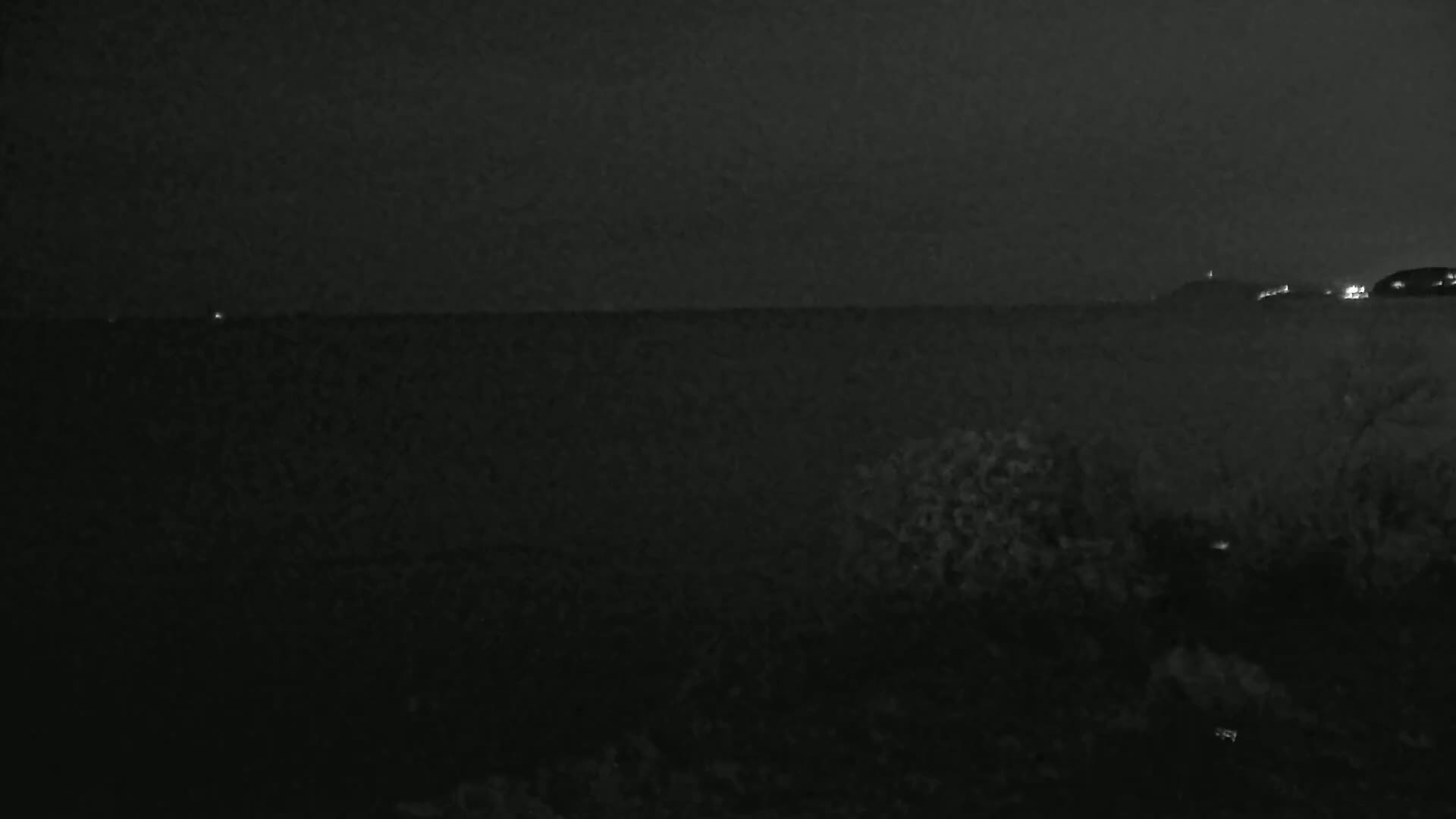 A dark, nighttime landscape shows a body of water with a distant, dimly lit shoreline under a cloudy sky.