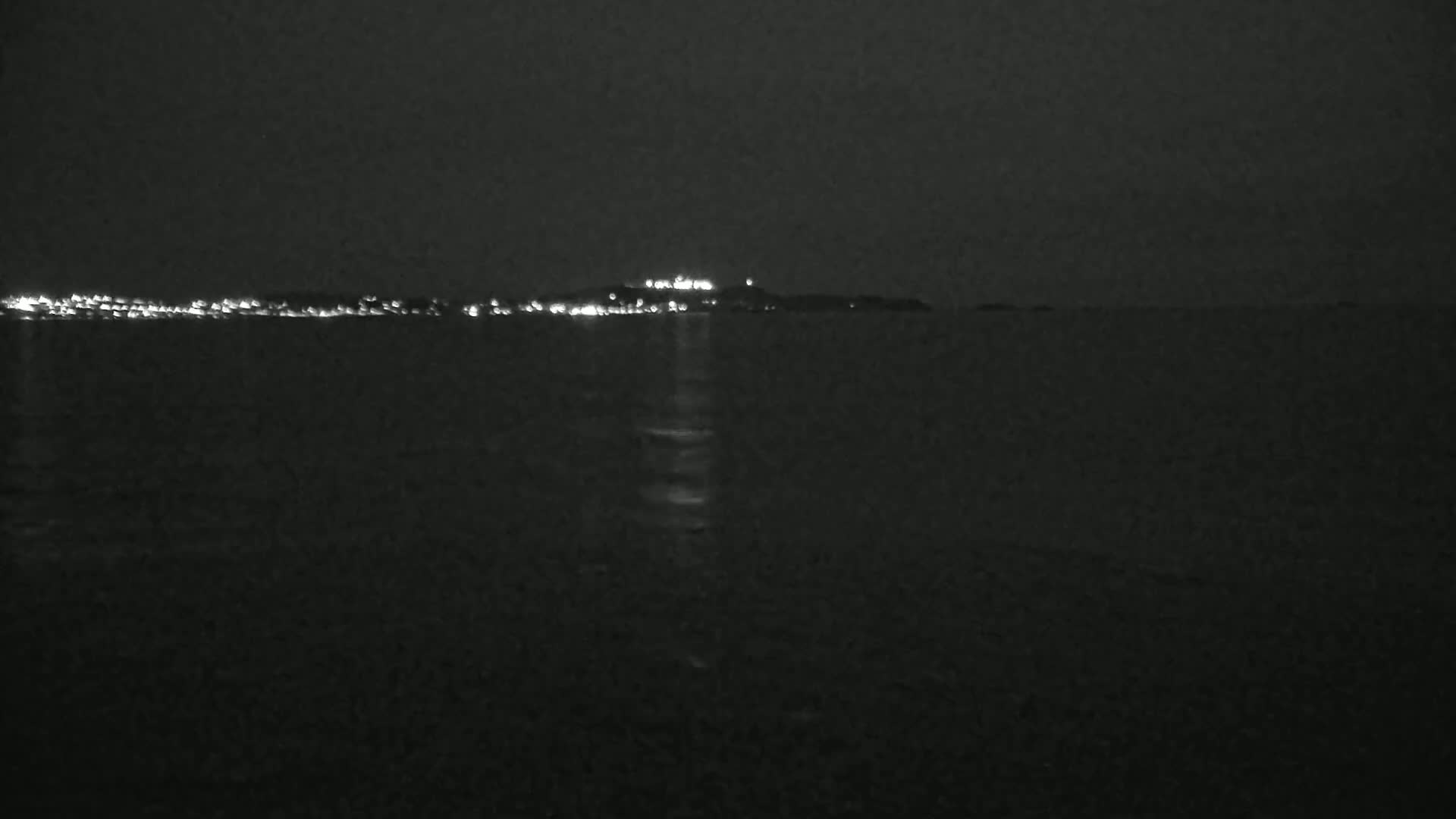 A dark, nighttime view shows a moonlit body of water reflecting lights from a distant, illuminated shoreline.
