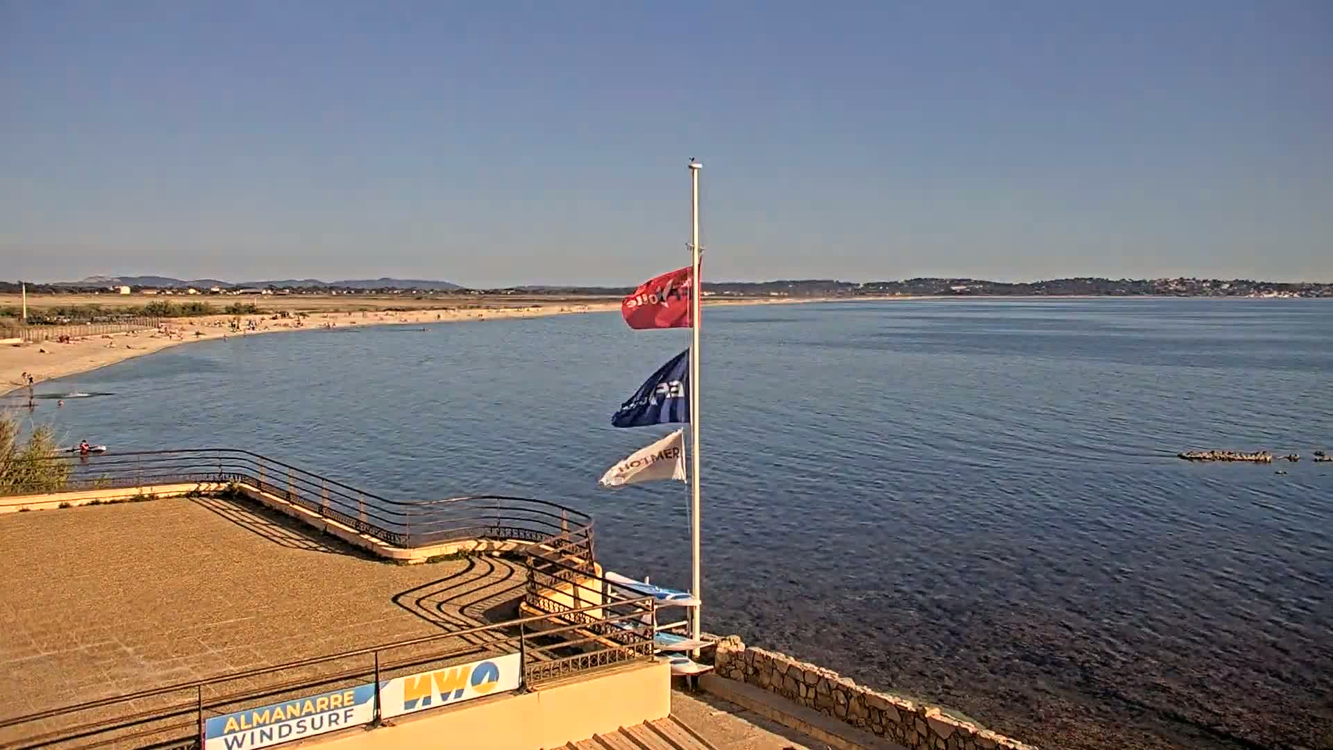 A sandy beach with people sunbathing is visible across a calm body of water from an elevated vantage point, three flags are mounted on a pole near a stone structure.