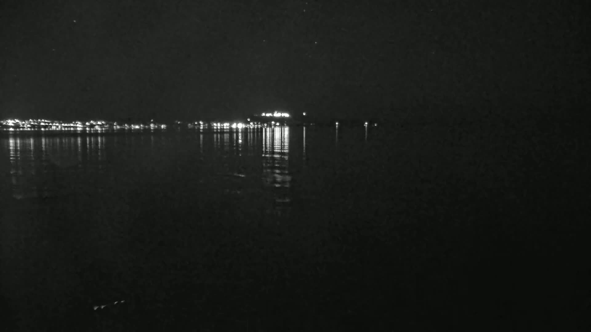 A dark body of water reflects the lights of a distant town at night under a clear sky.