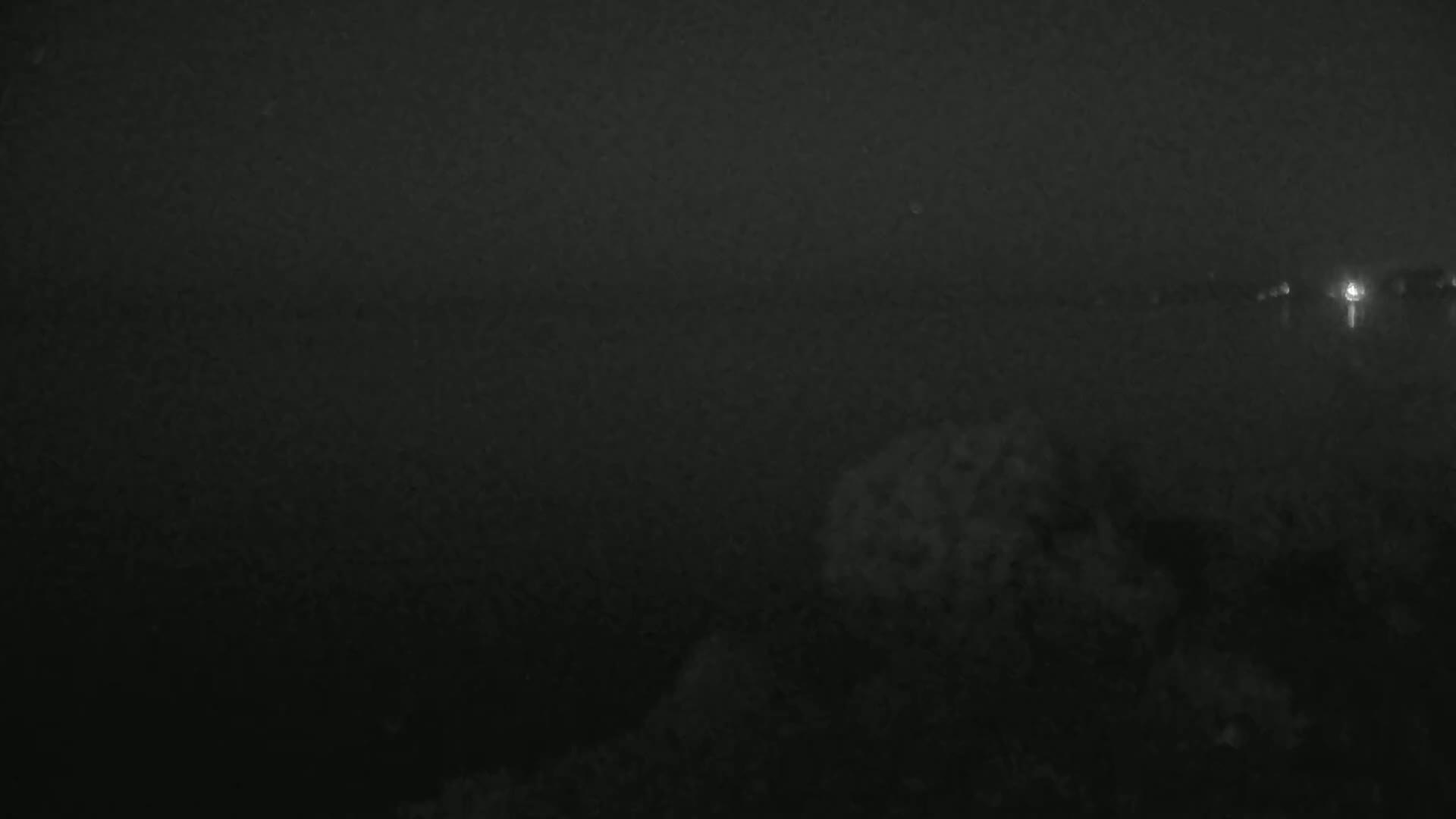 A dark, nighttime image shows a body of water under a hazy sky, with faintly visible lights in the distance.