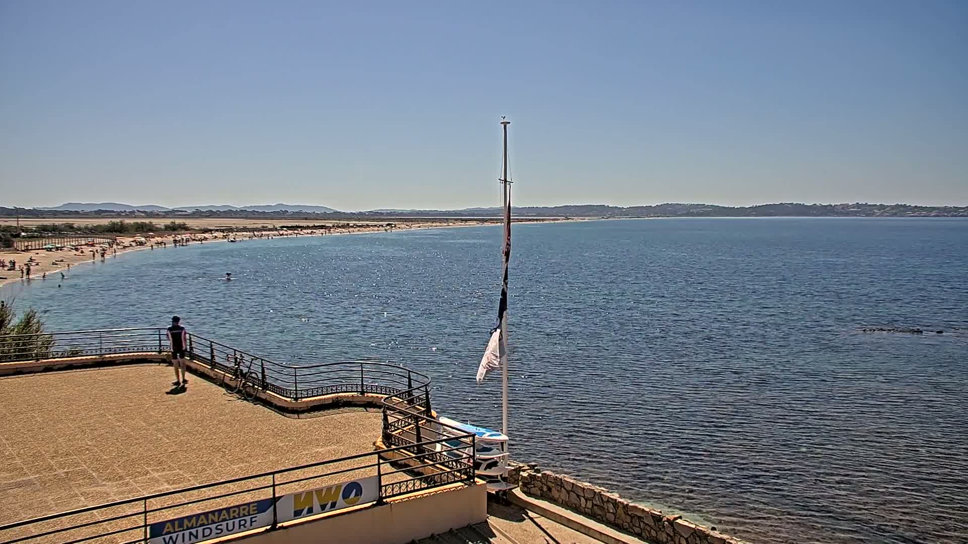 A long sandy beach stretches along a calm, blue sea under a clear, sunny sky, viewed from an elevated vantage point with a person standing on a nearby patio.