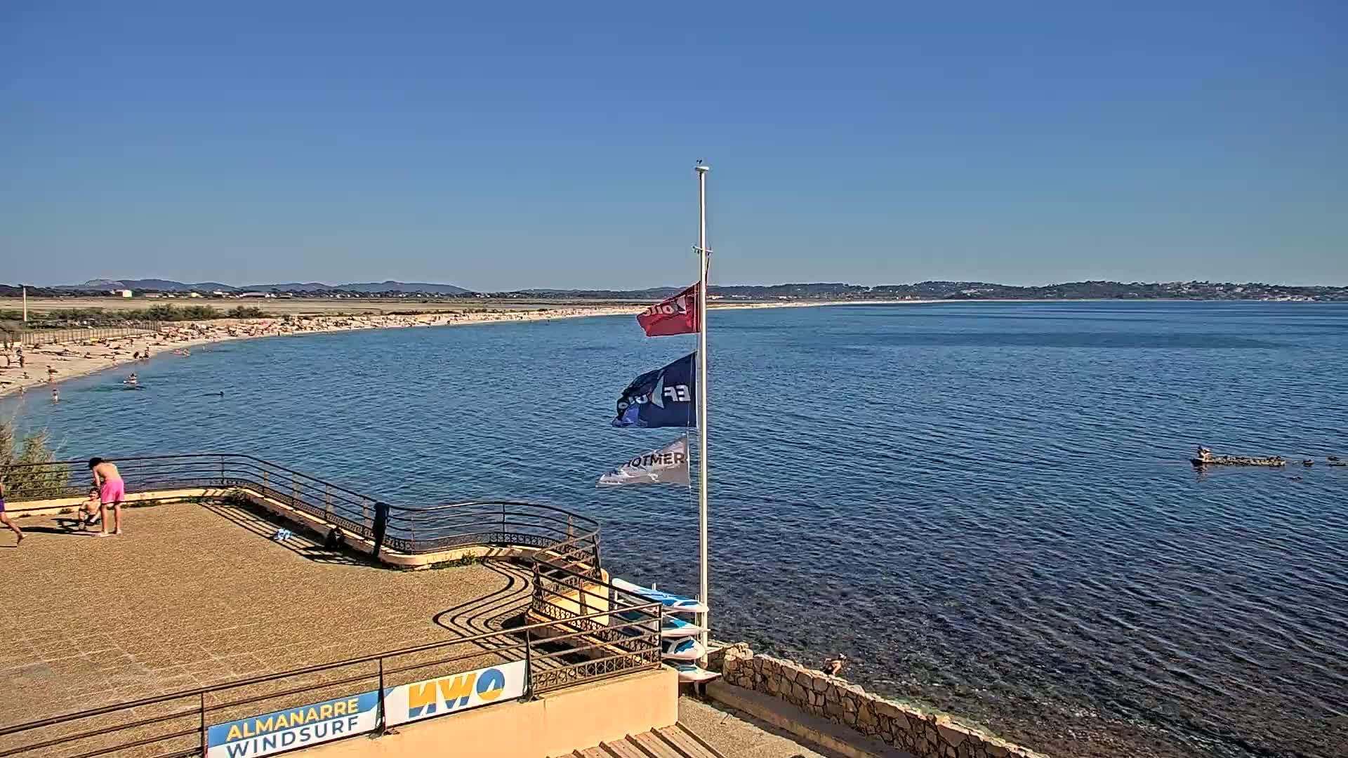 A sandy beach with many people is visible, adjacent to calm blue water under a clear blue sky; a paved area with a railing is in the foreground.