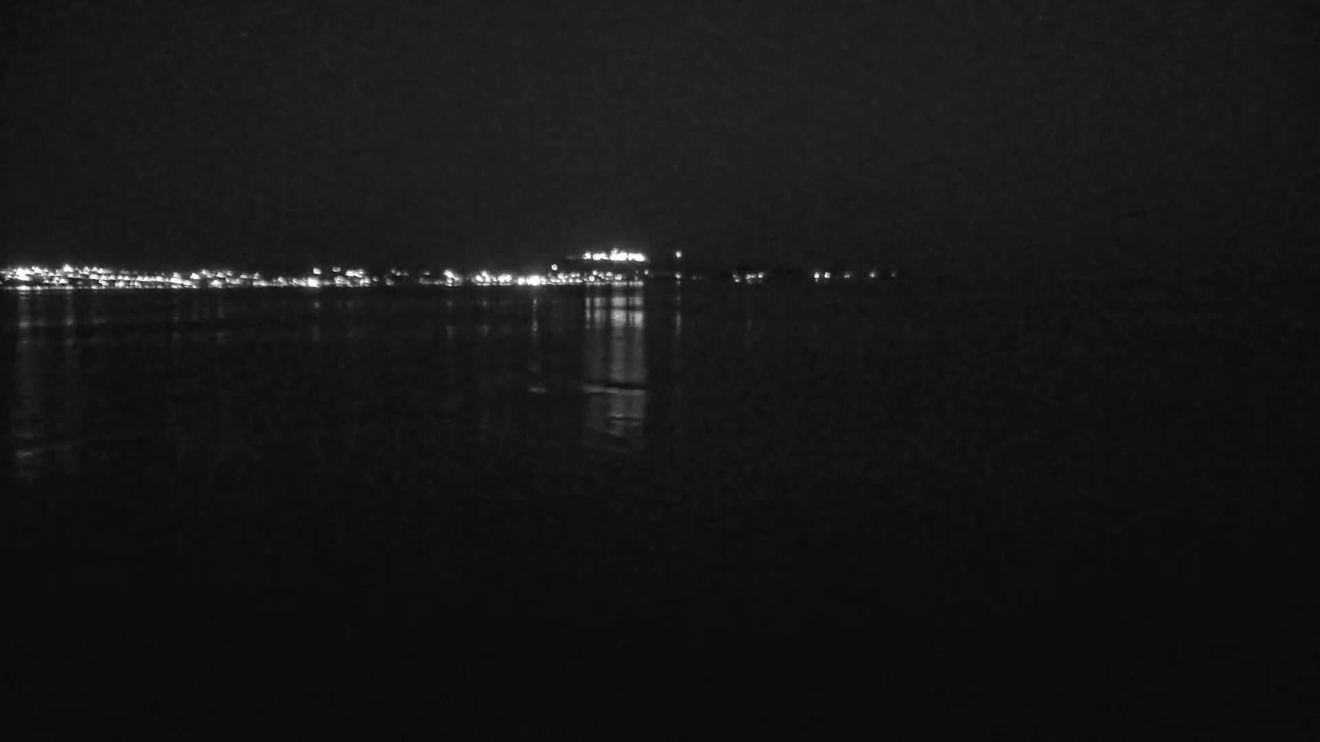 A dark, nighttime scene shows a body of water reflecting the lights of a distant city skyline.