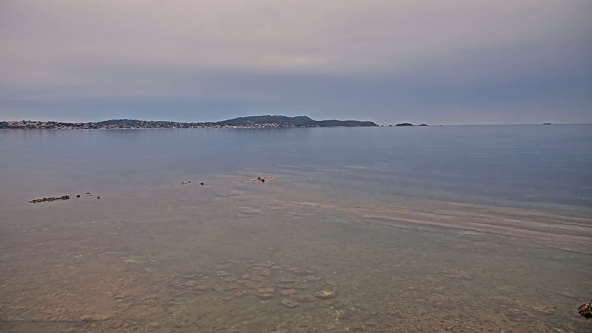 Under a cloudy sky, clear shallow water reveals a rocky seabed extending to a distant island and shoreline.