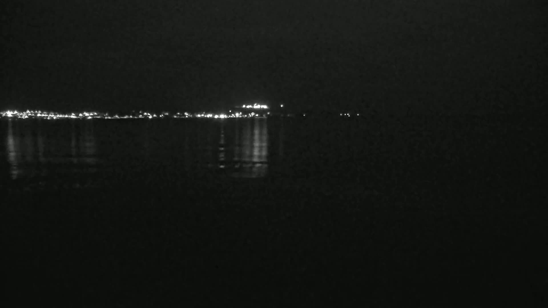 At night, city lights illuminate a dark body of water, reflecting faintly on its surface.