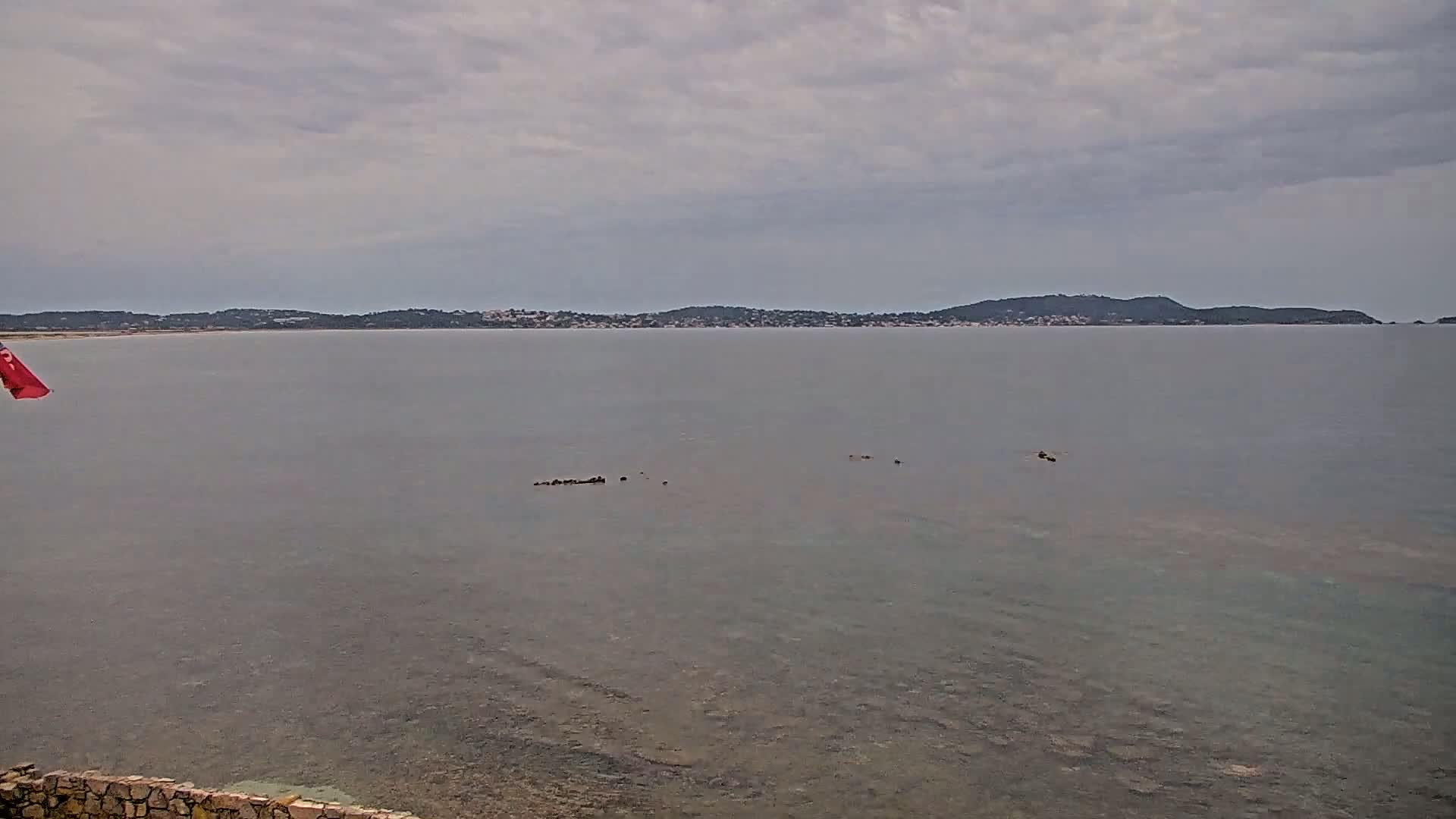 A calm, overcast day reveals a body of water with a distant shoreline featuring low hills and a red flag partially visible in the bottom left corner.
