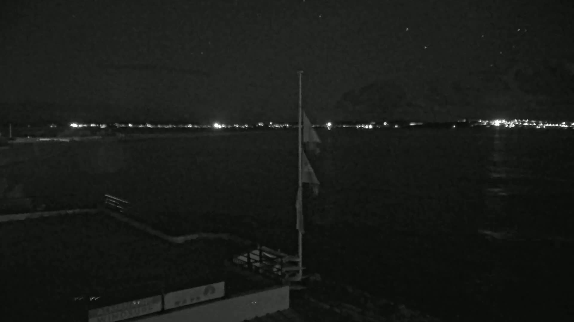 A nighttime view shows a flagpole with flags, a dark body of water, and a distant, faintly lit shoreline under a clear, starry sky.