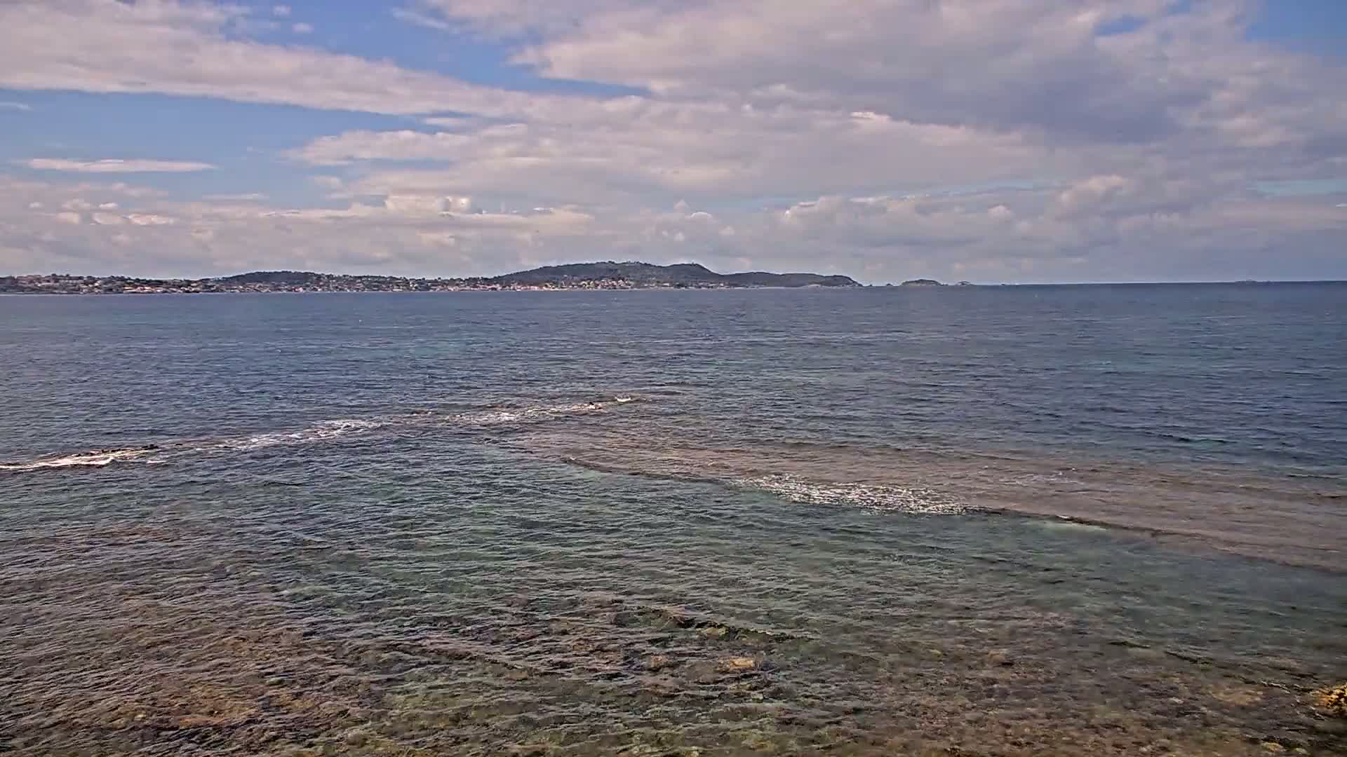 A partly cloudy sky overlooks a calm sea with a rocky shoreline in the foreground and a distant landmass on the horizon.