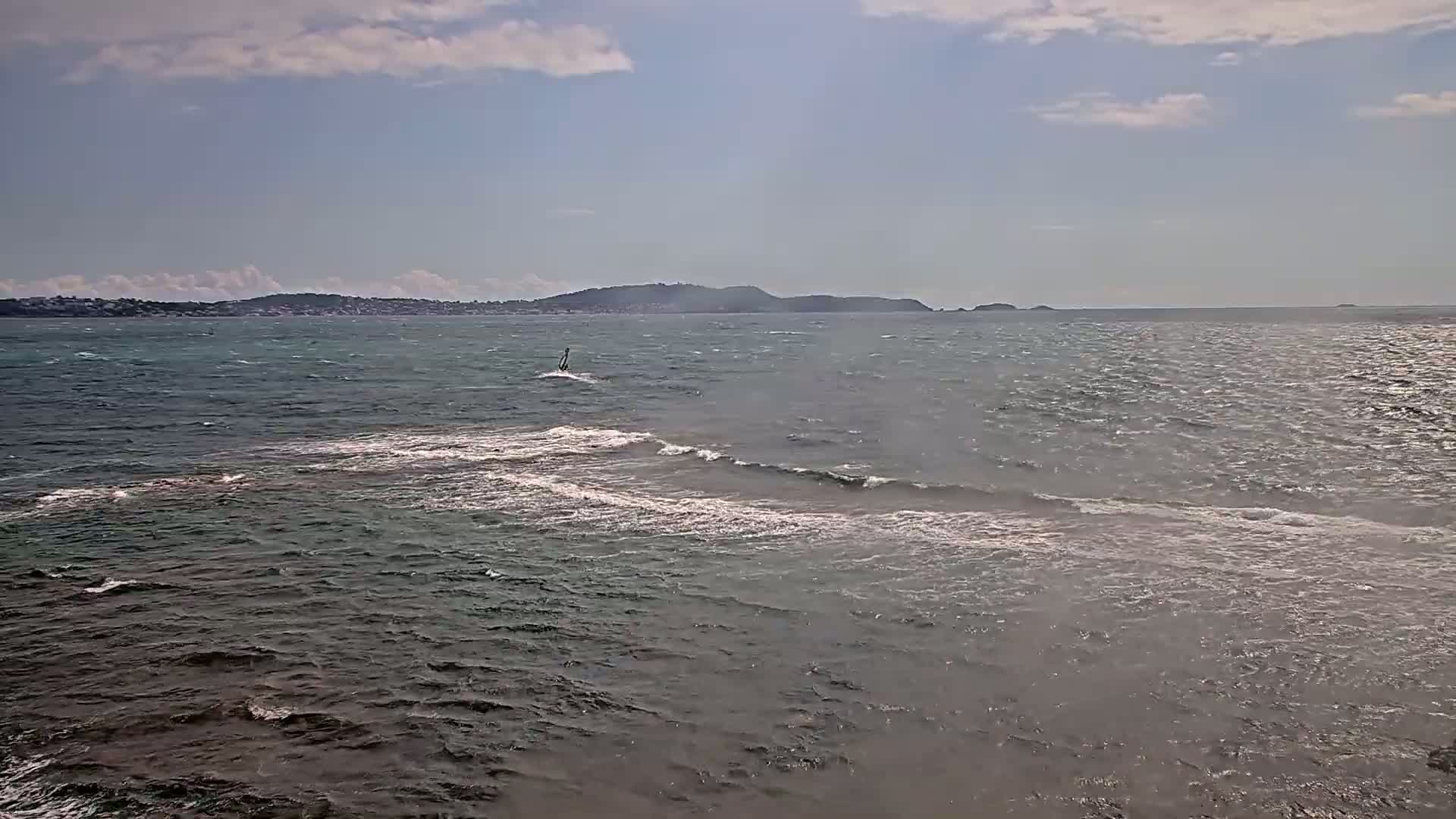 A windsurfer sails on a choppy sea under a partly cloudy sky, with a distant shoreline visible.