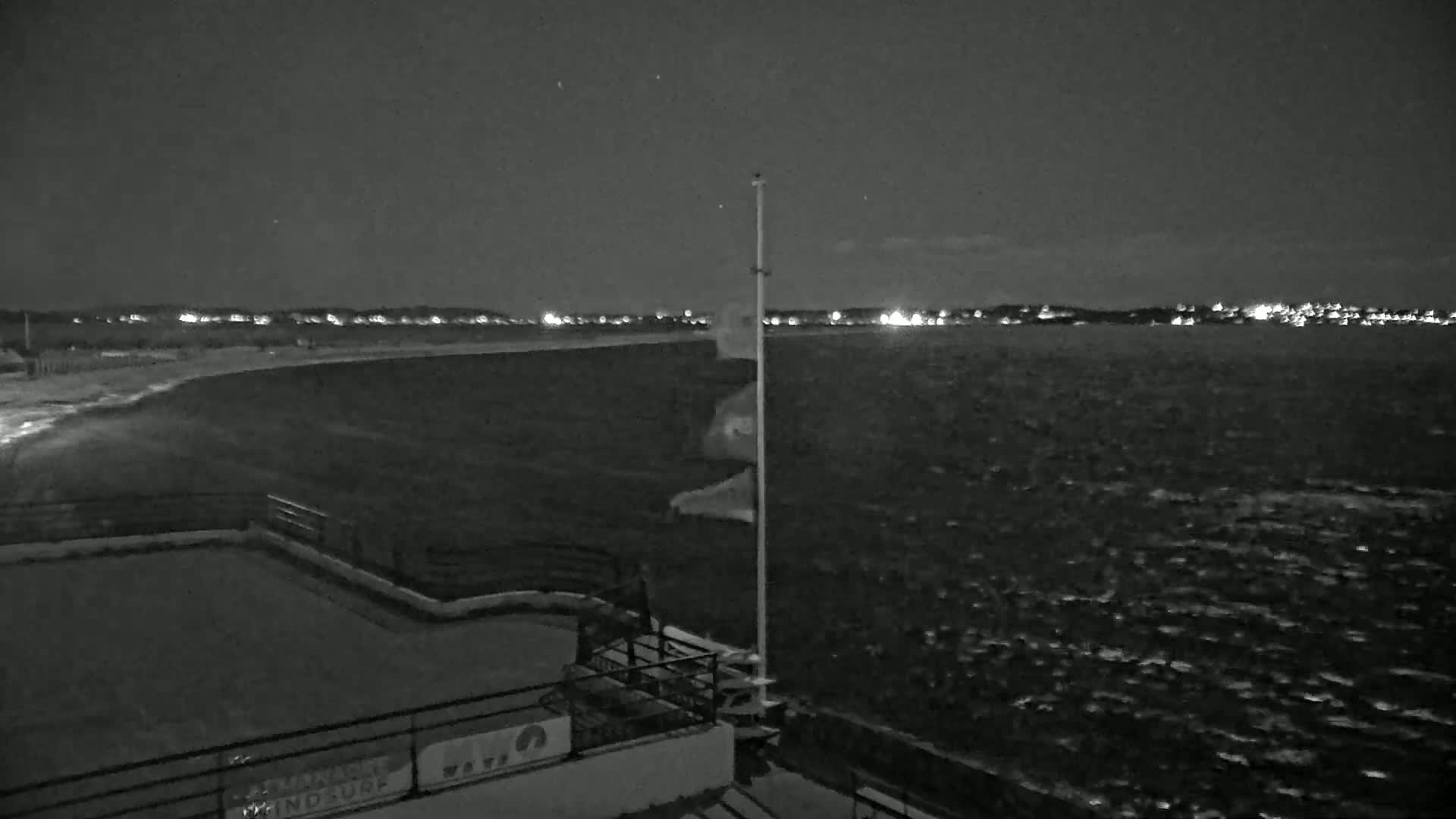 A nighttime view of a dark, wavy body of water with a shoreline featuring lights in the distance, and a flagpole with flags on it in the foreground.