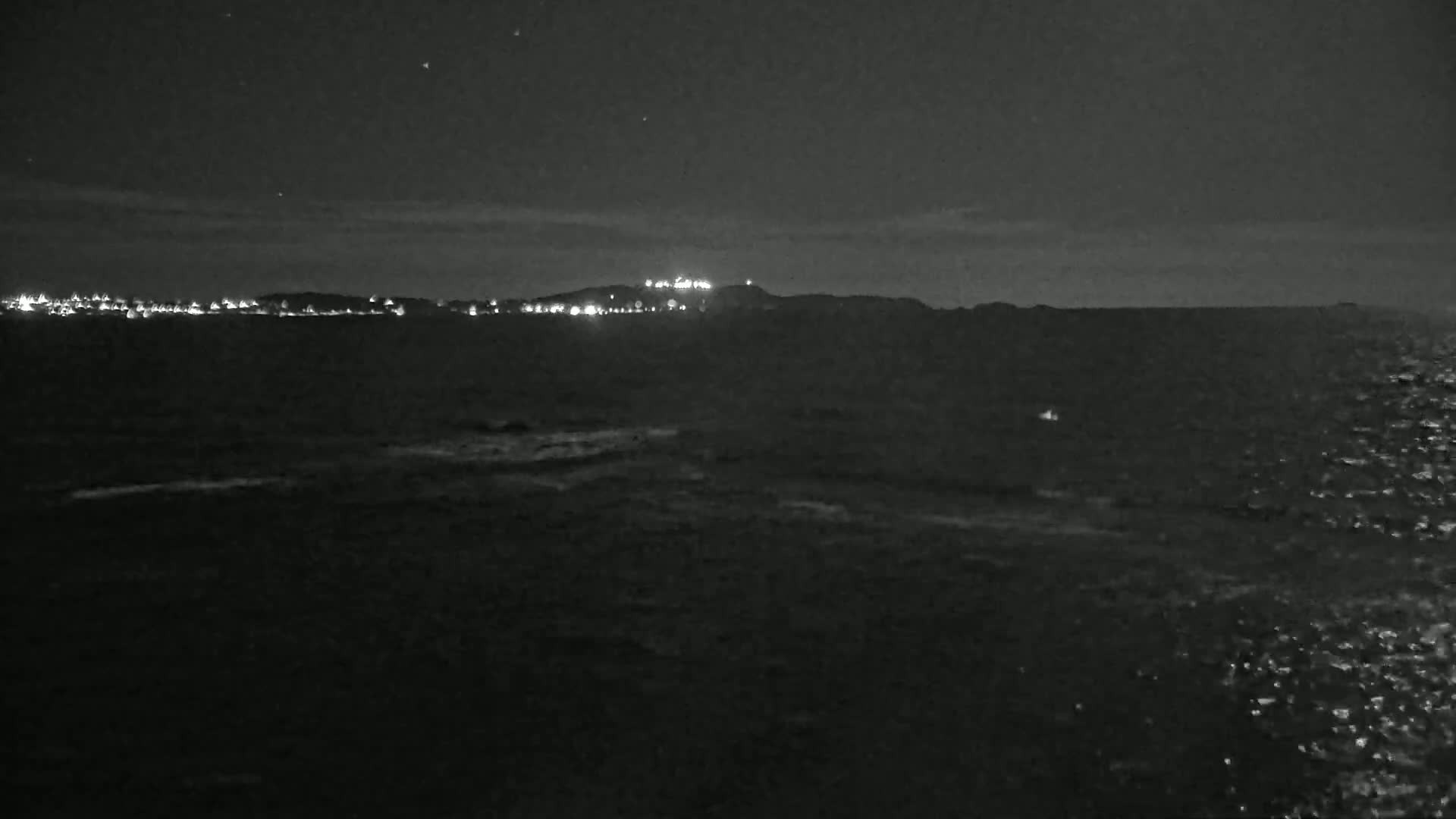 A dark, nighttime seascape shows a distant, lit shoreline and hill under a mostly clear sky with a gently rippling sea in the foreground.