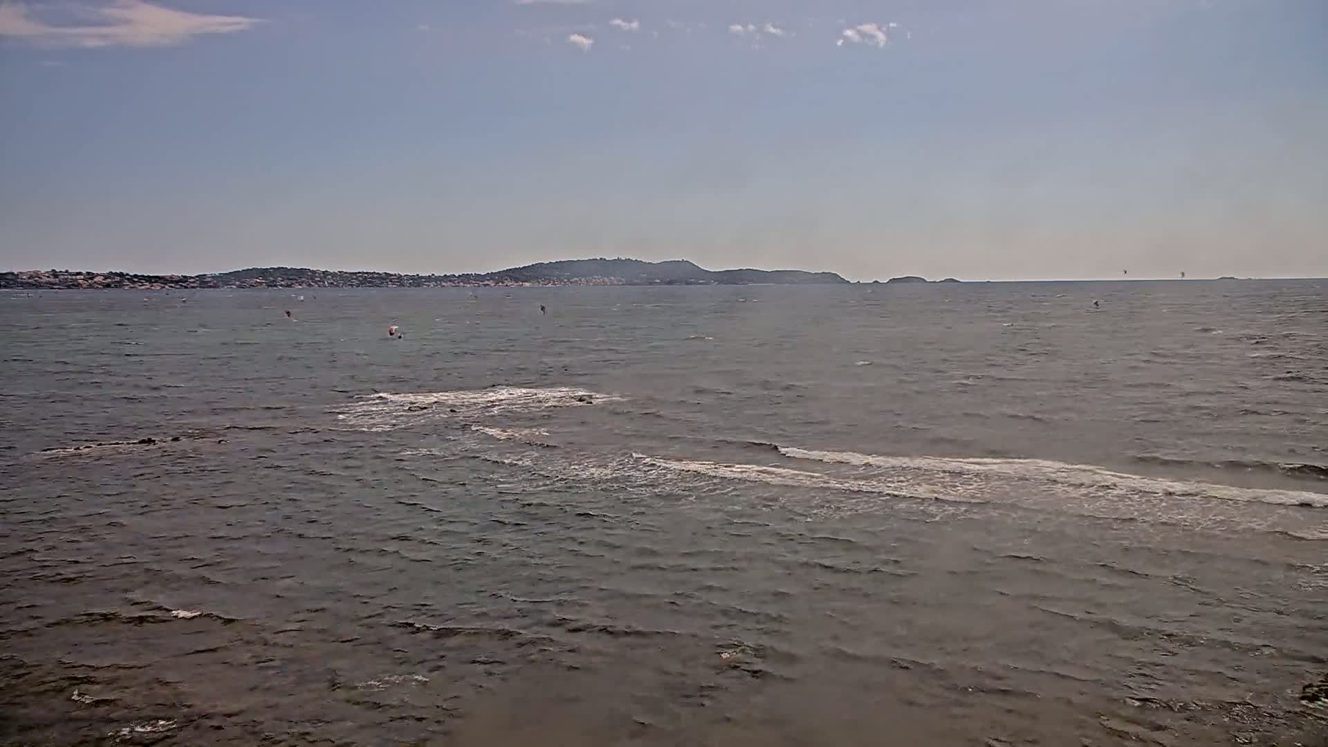 A mostly calm sea with small waves stretches to a distant shoreline under a partly cloudy sky, with a few windsurfers visible on the water.