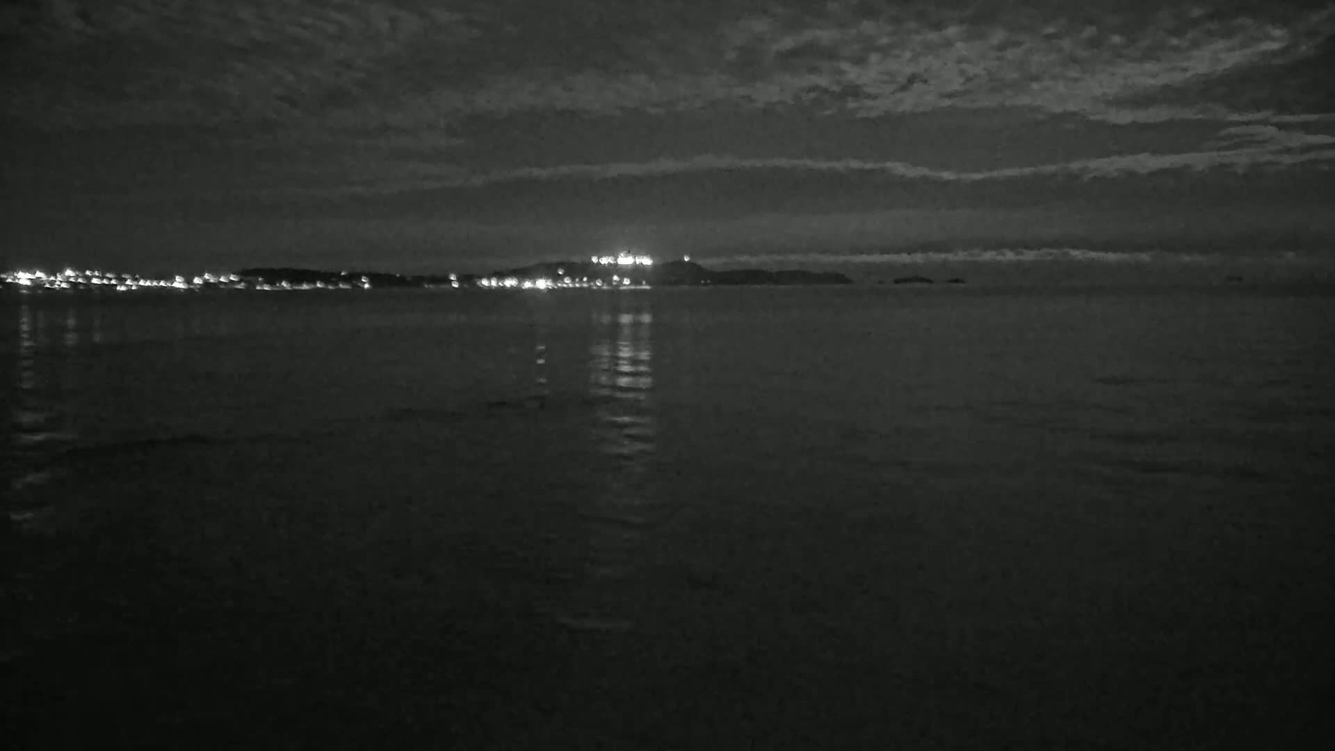 A dark, nighttime scene shows a calm body of water reflecting the lights of a distant city on a cloudy night.