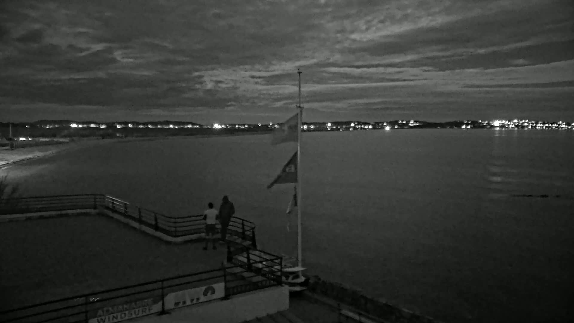 Two people stand on a platform overlooking a calm body of water at night, under a cloudy sky, with lights visible on the far shore.