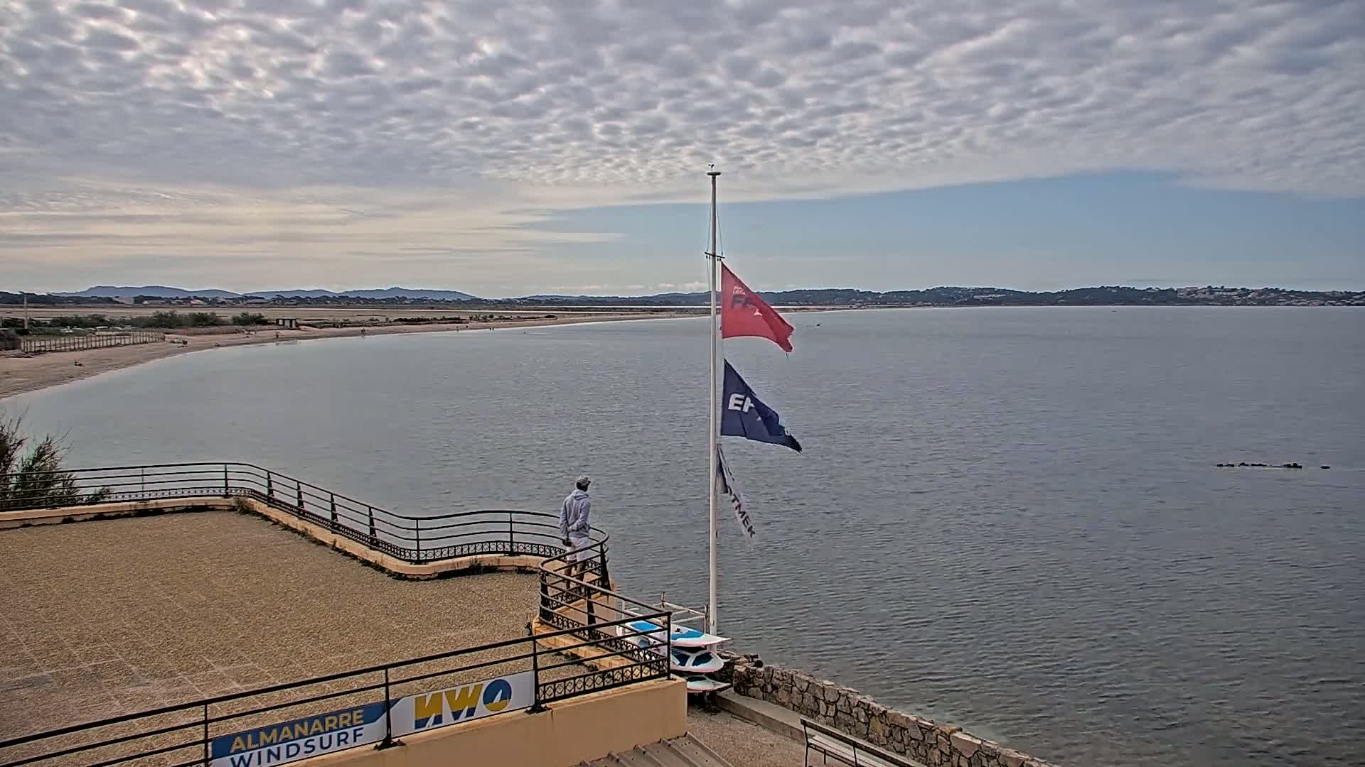 A person stands on a balcony overlooking a calm bay with a sandy beach and distant hills under a cloudy sky.