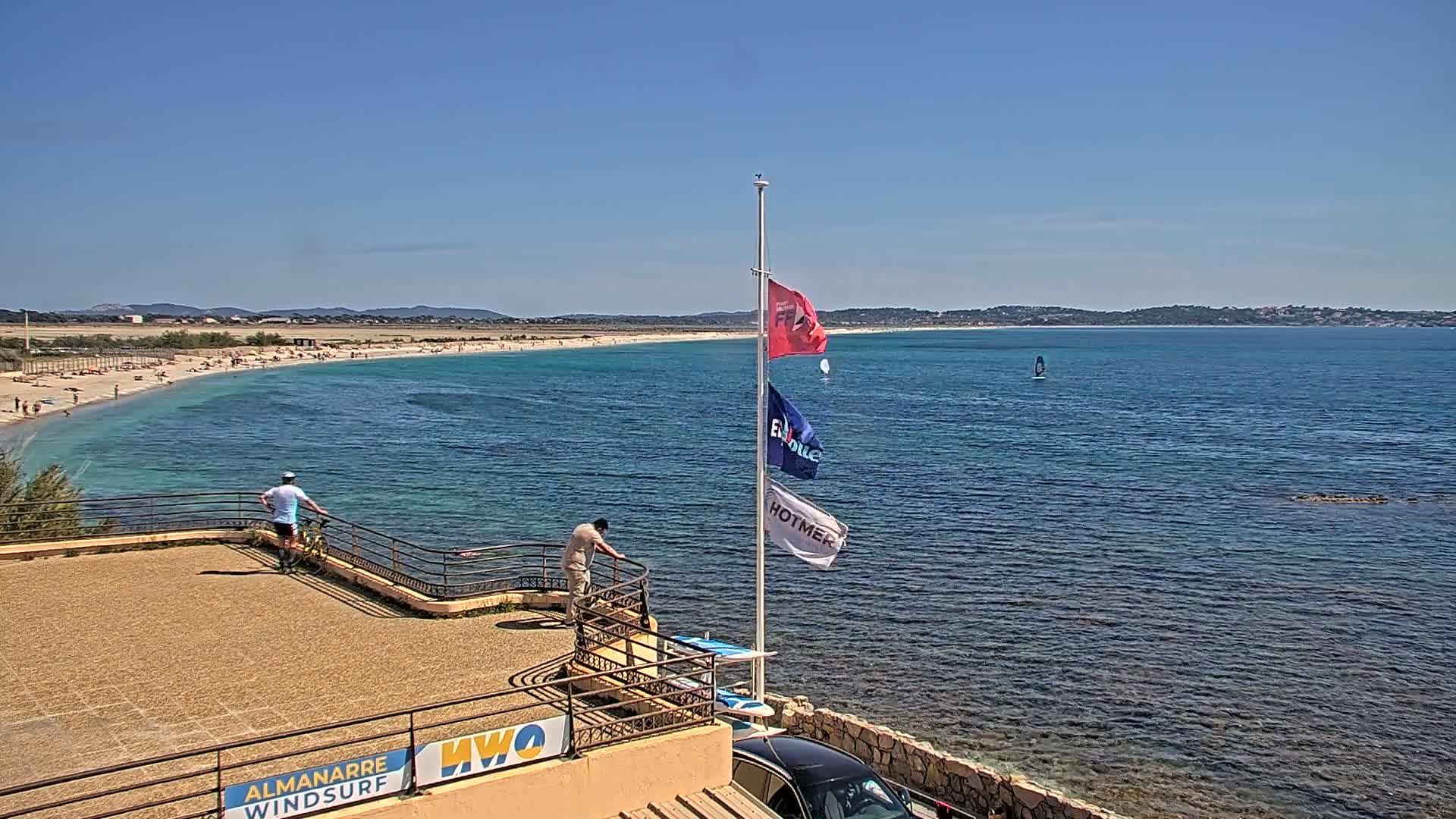 A sunny day shows a calm ocean with a sandy beach filled with people, viewed from an elevated vantage point with a few people and windsurfing equipment nearby.