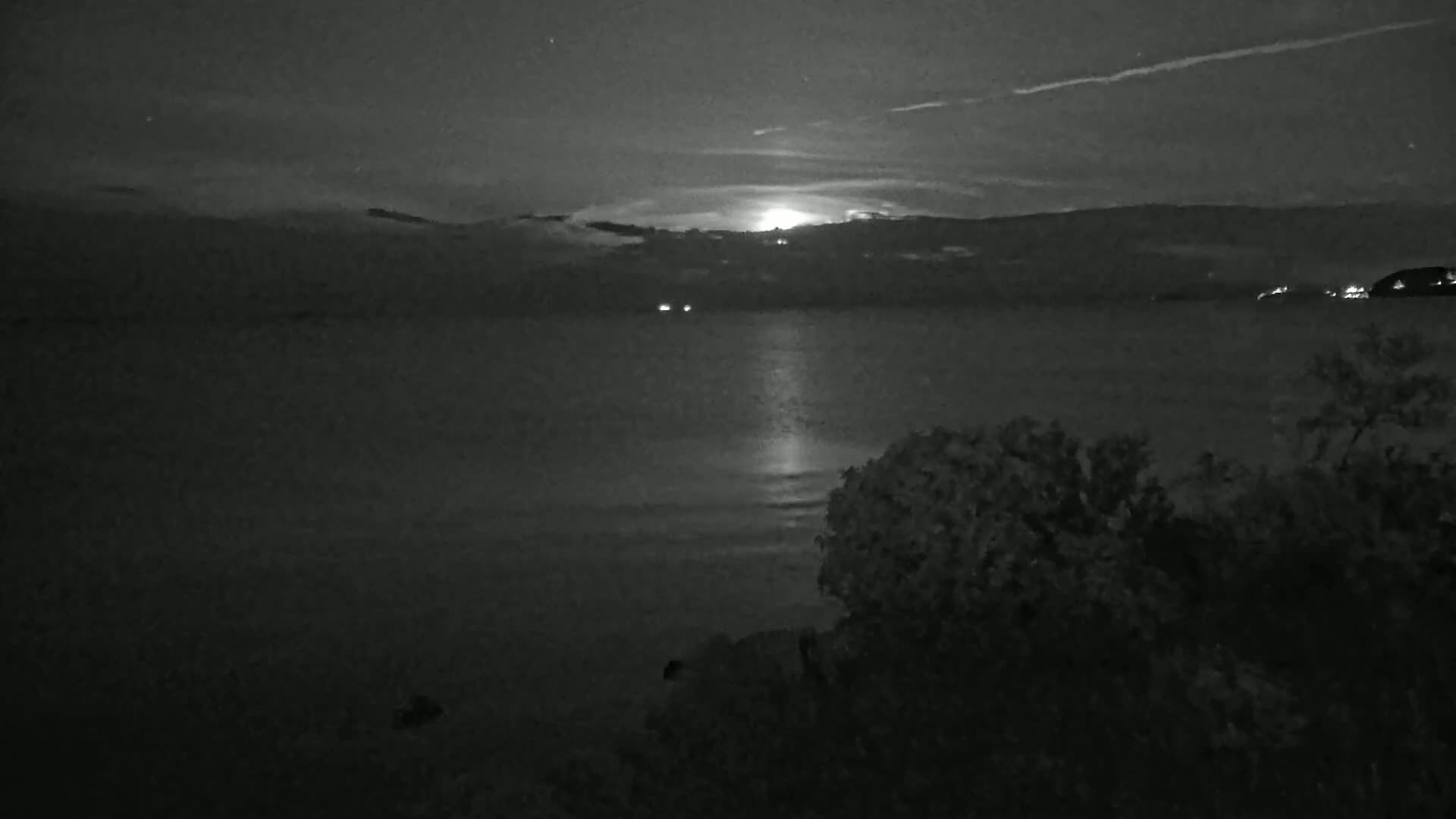 A moonlit nighttime scene shows a calm body of water reflecting the light, with dark land and vegetation in the foreground and background under a partly cloudy sky.