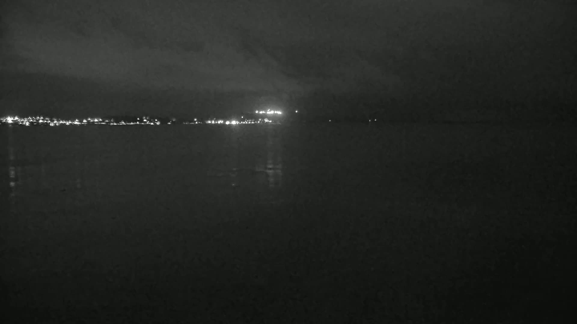 A dark, nighttime scene shows a body of water reflecting the lights of a distant city under a cloudy sky.