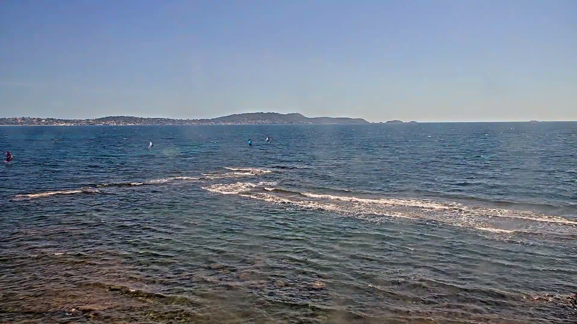 A calm, sunny day reveals a coastal scene with several windsurfers on the water near a rocky shoreline and a distant landmass.
