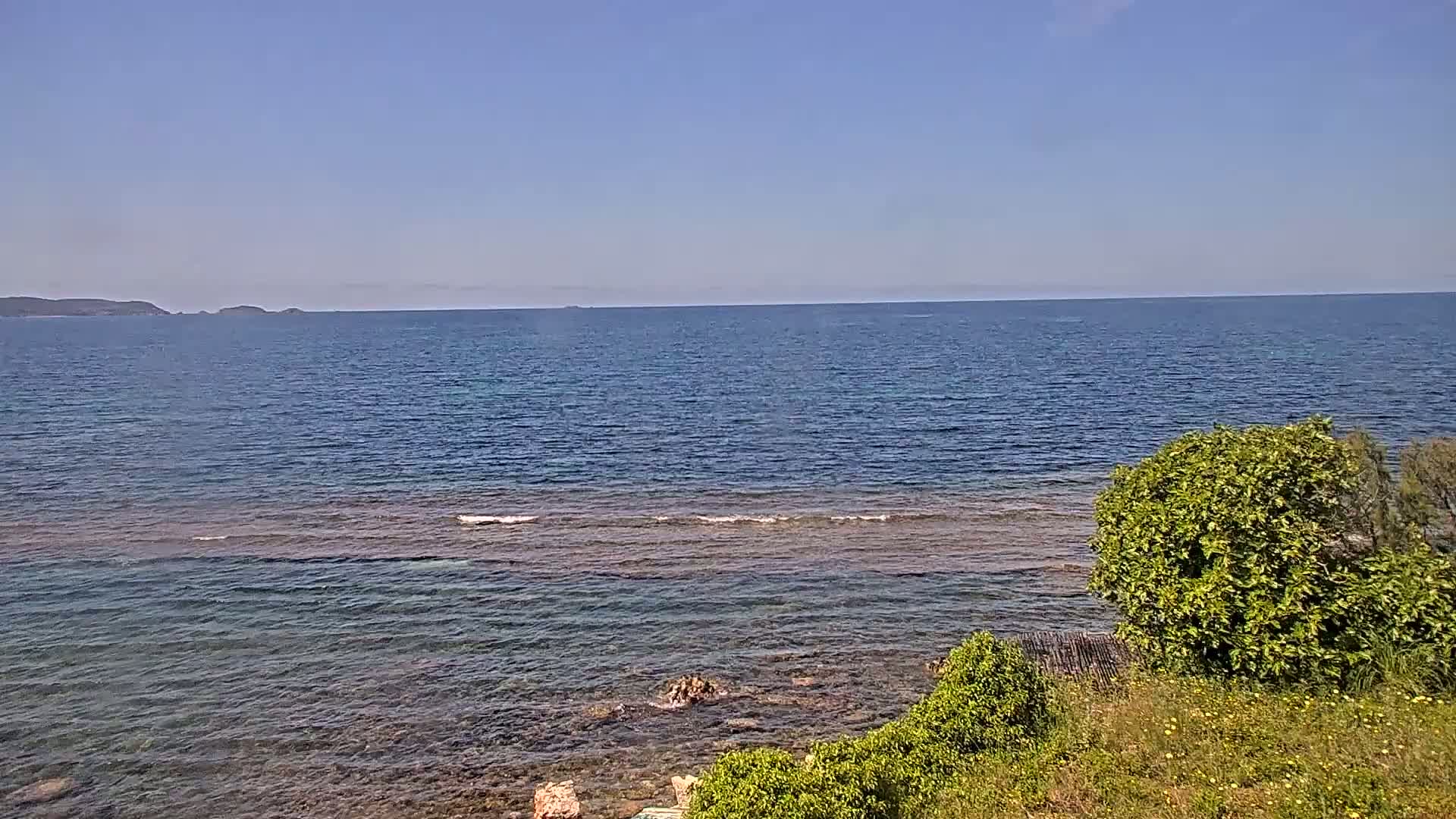 A calm, clear blue sea stretches to a distant, low-lying landmass under a bright blue sky, with green vegetation in the foreground.
