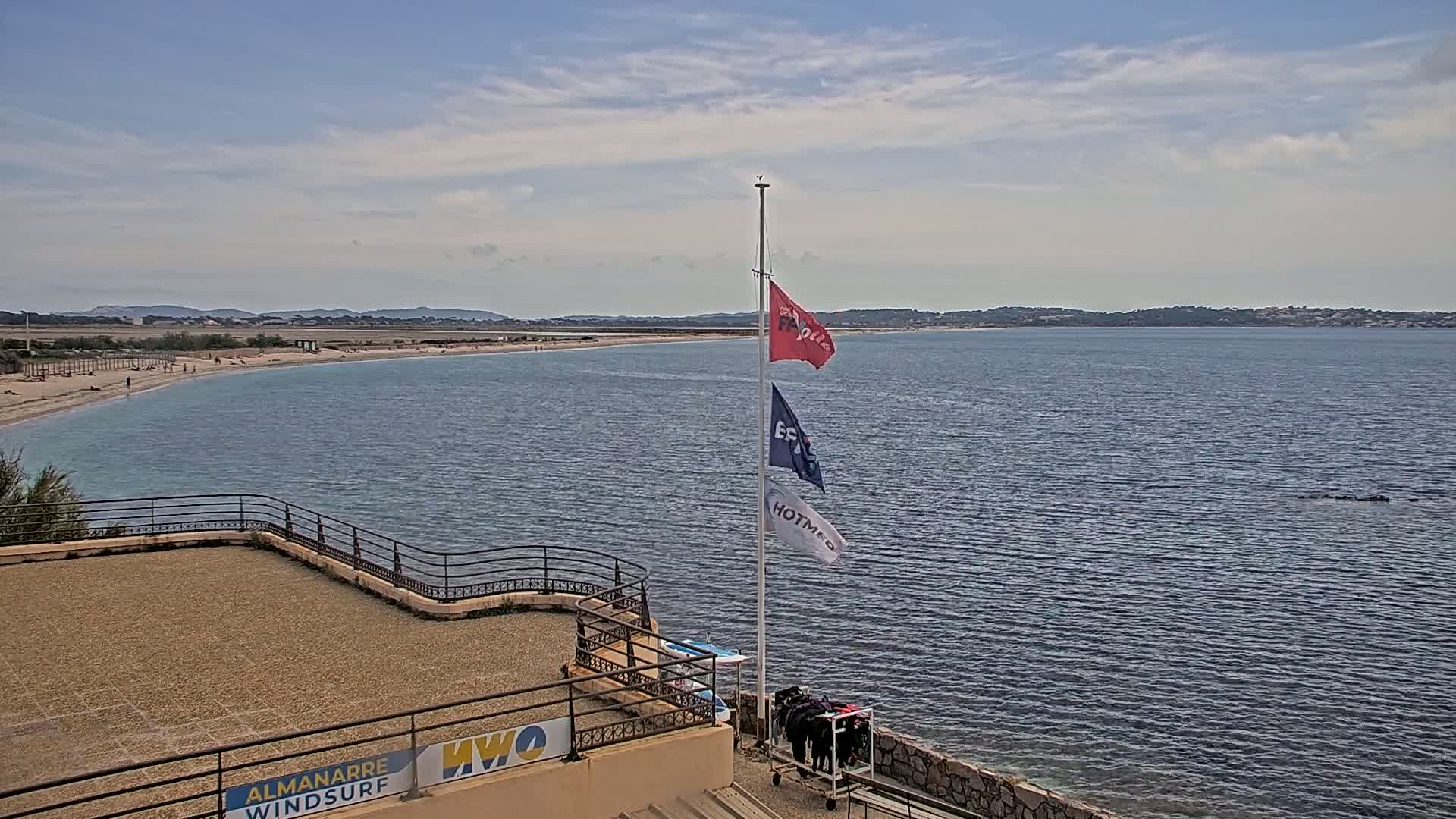 A sandy beach stretches along a calm bay under a partly cloudy sky, with several flags on a pole near a stone building.