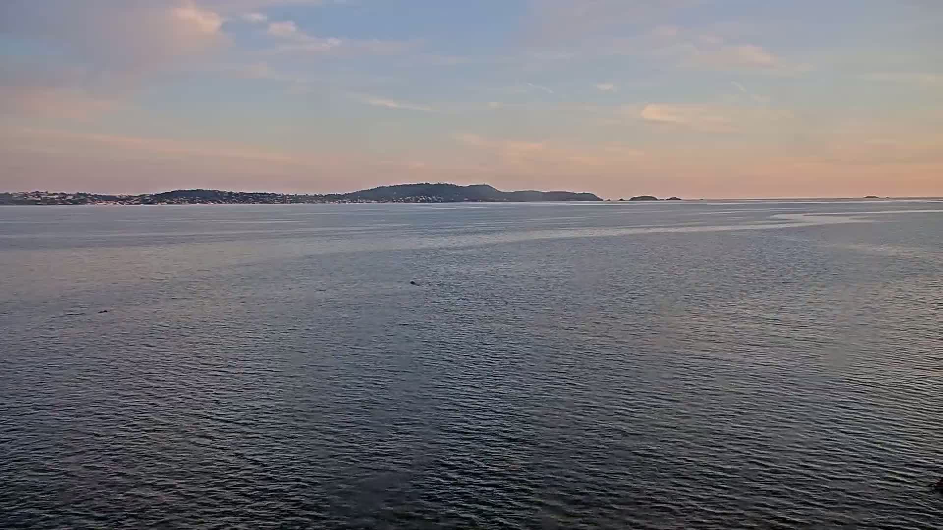 A calm, dark-blue body of water stretches to a distant shoreline of low-lying hills under a pale, cloudy sunset sky.