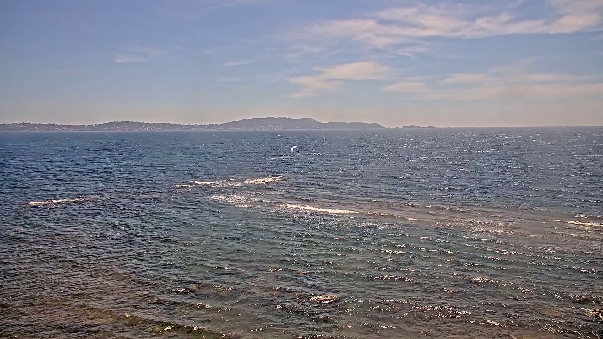 A person kitesurfs on a partly sunny day over a calm sea with a distant landmass visible on the horizon.