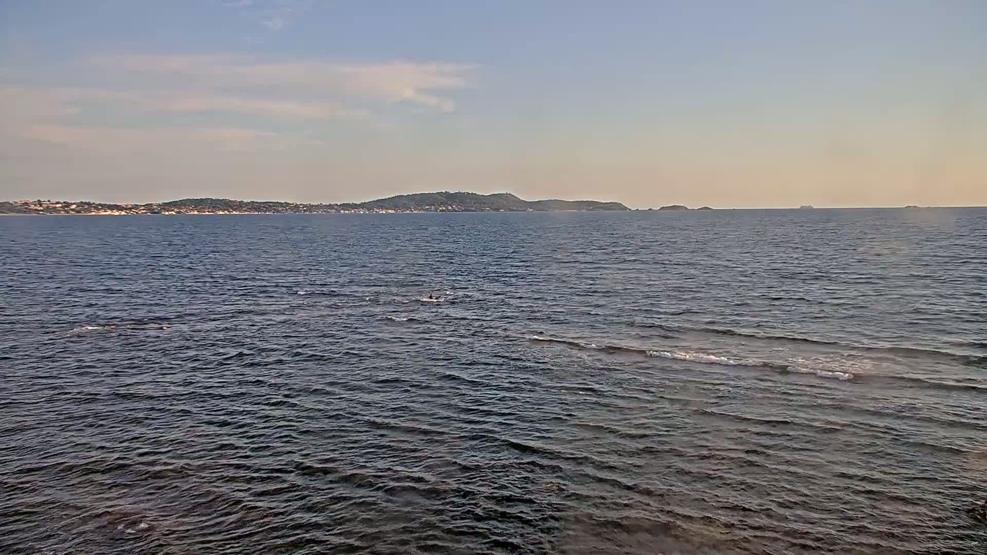 A calm, dark-blue sea stretches to a distant coastline under a pale-blue, mostly clear sky.